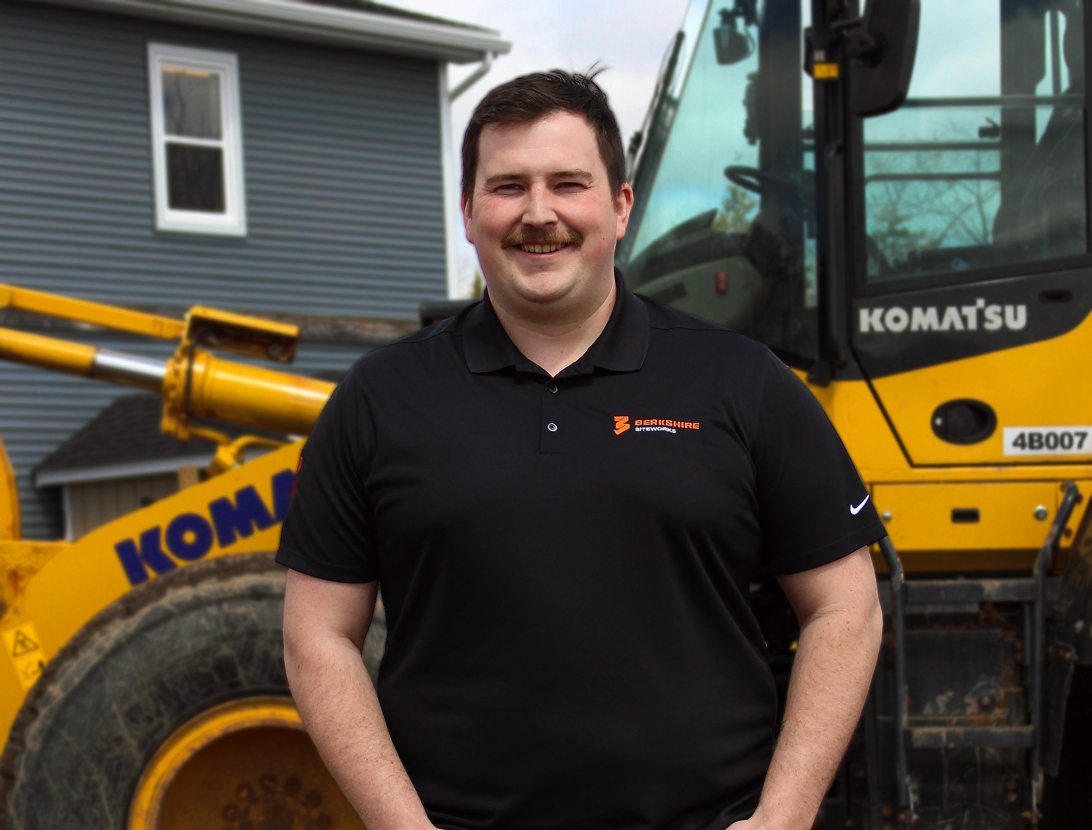 Man smiling in front of construction equipment and a house, wearing a black polo shirt with logos.