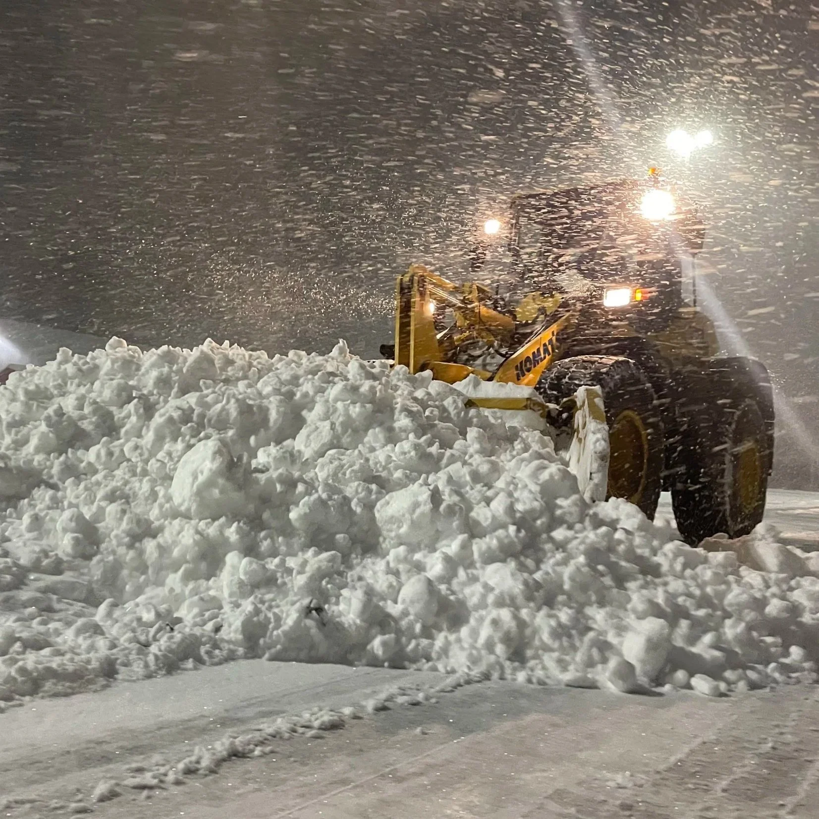A snowplow clearing snow from a road during a snowstorm at night, with snow flying in the air and headlights illuminating the scene.