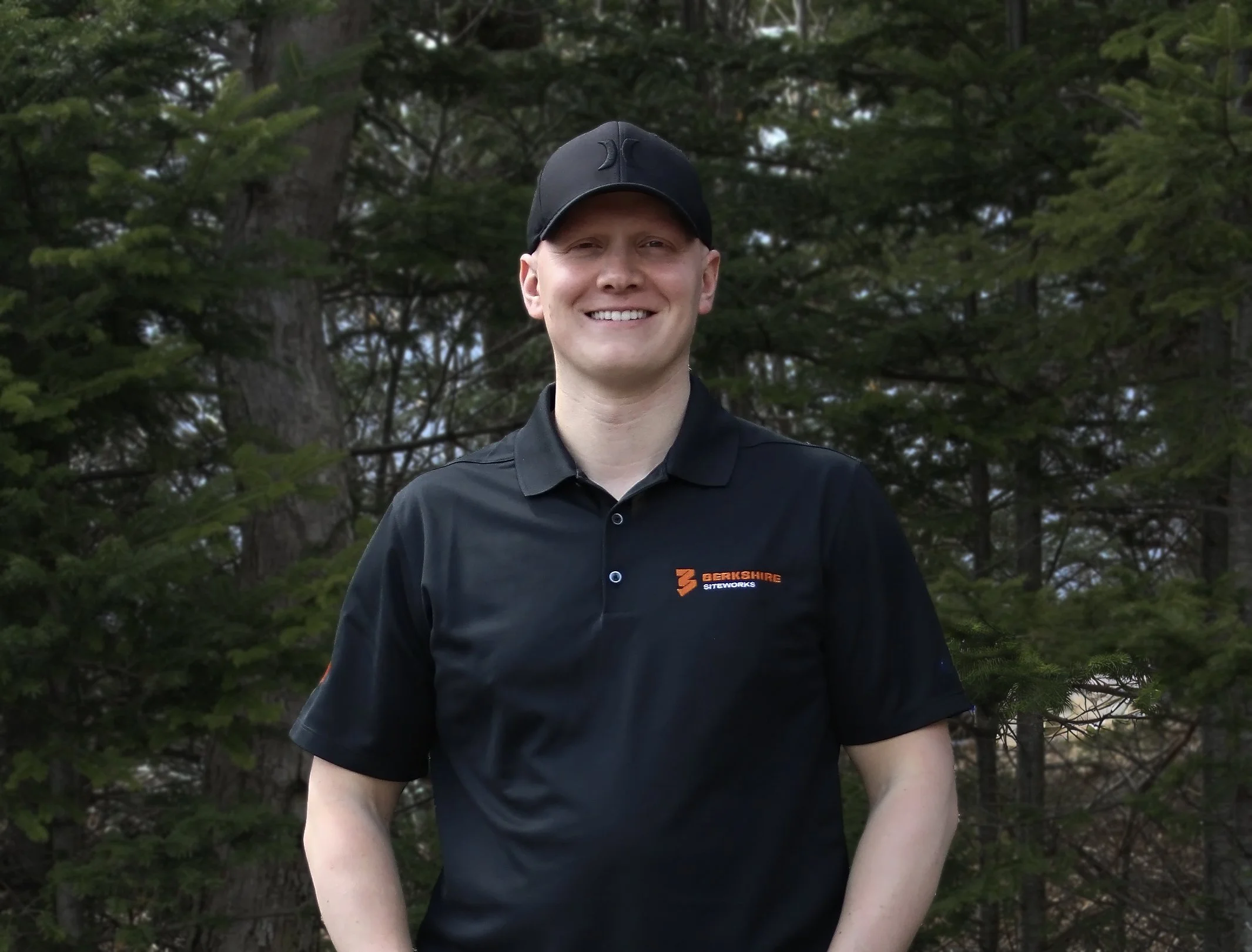 A young man smiling outdoors wearing a black cap and black polo shirt with orange and white logo on the chest, standing in front of green trees.