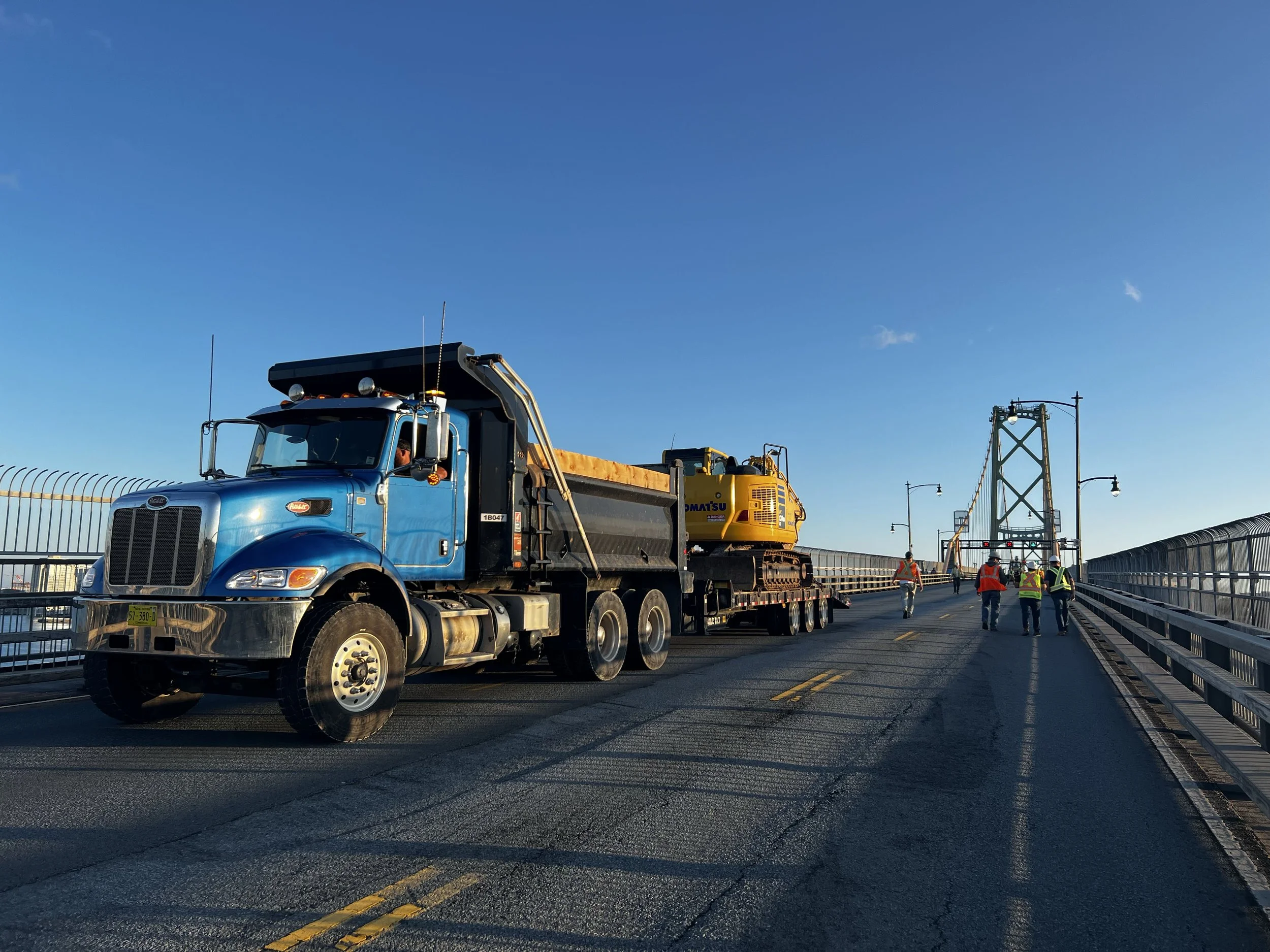 Construction workers walking on a bridge with a large truck and heavy equipment, blue sky in the background.