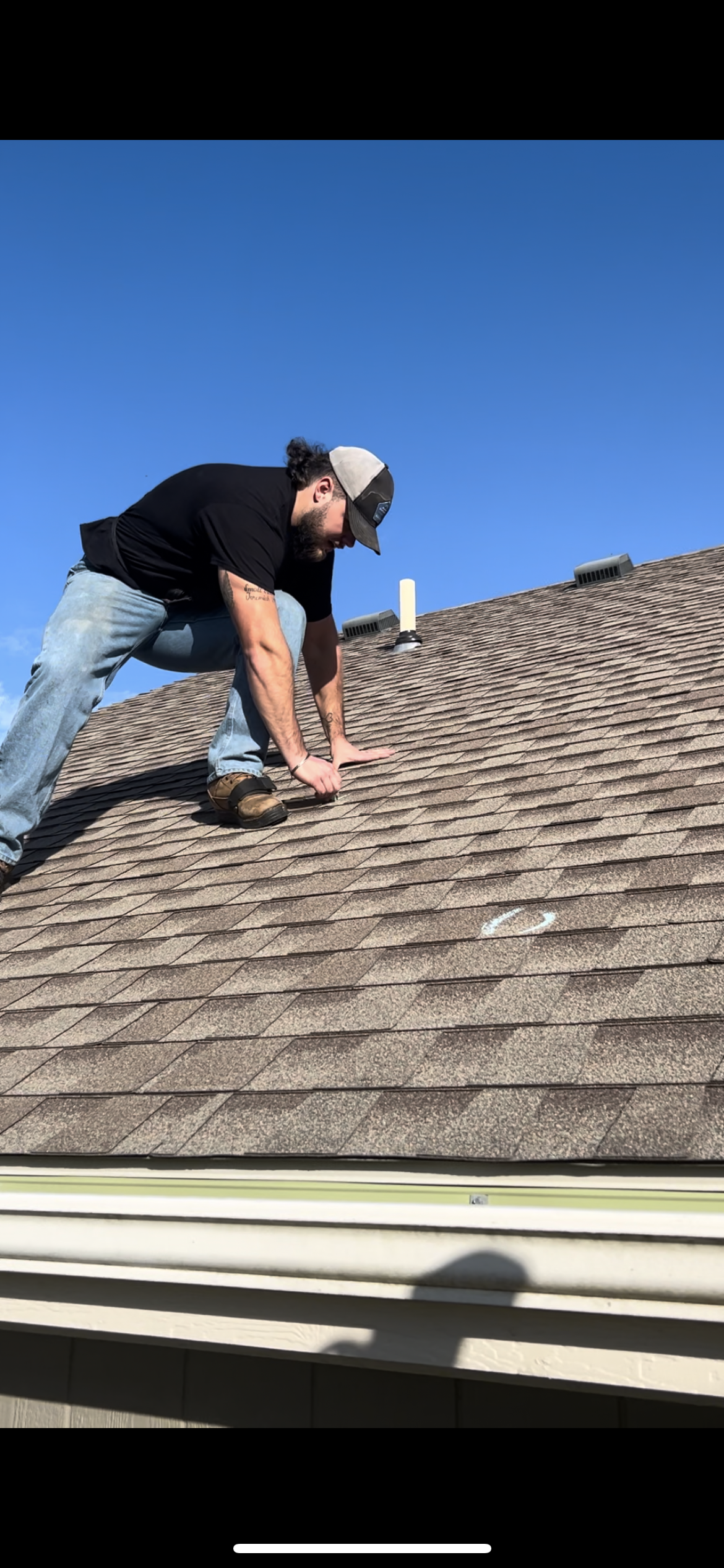 Man inspecting and working on a residential roof with brown shingles, under a clear blue sky.