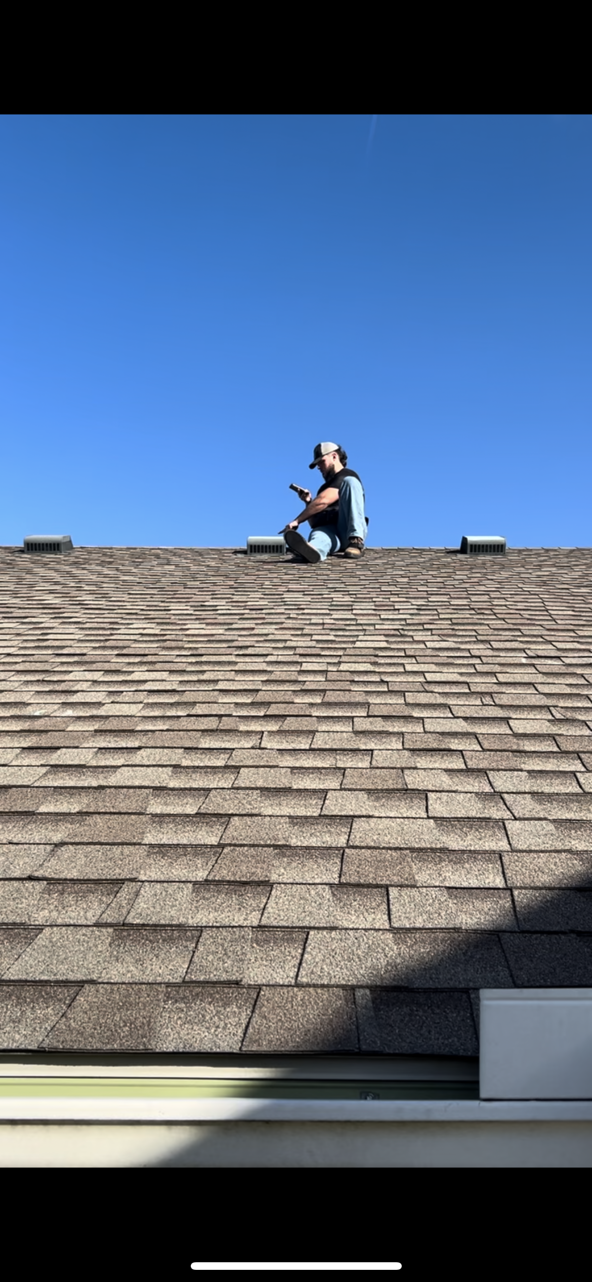 A man sitting on a rooftop shingle, looking at his phone, with a blue sky above.