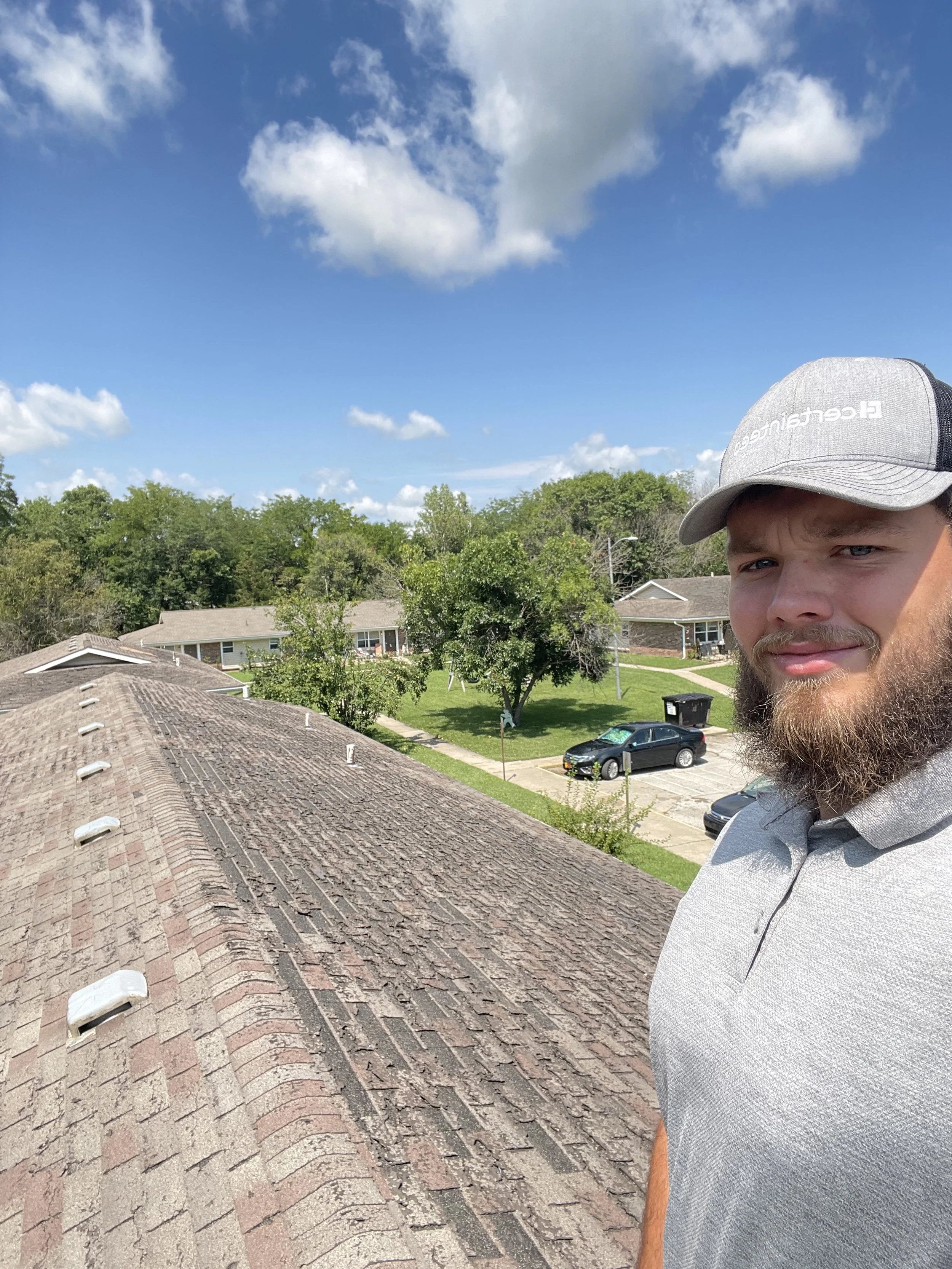 A man wearing a gray cap and gray polo shirt taking a selfie on a residential rooftop. The background shows a suburban neighborhood with trees, parked cars, houses, and a bright blue sky with scattered clouds.
