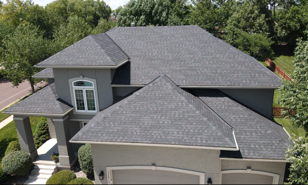 Aerial view of a two-story house with a gray roof, gray exterior walls, a front porch with steps, surrounded by shrubs and trees, in a suburban neighborhood.
