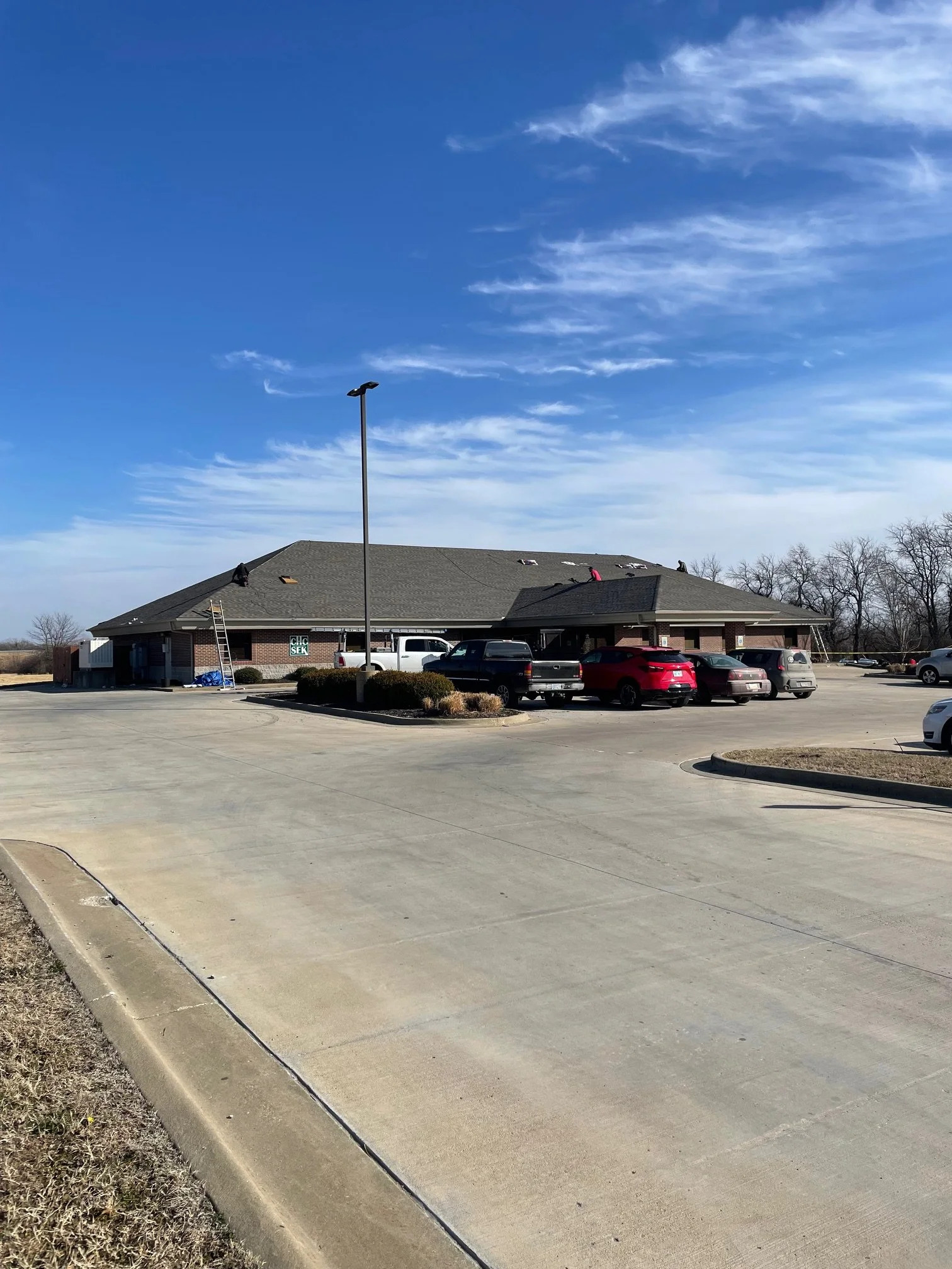 A parking lot with a single-story brick building under a blue sky with some clouds. Several cars are parked, and people are on the roof of the building working on it.