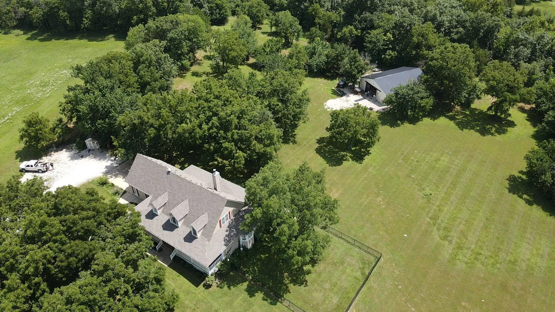 Aerial view of a large house surrounded by trees and open grassy areas, with a gravel driveway and a white pickup truck nearby.