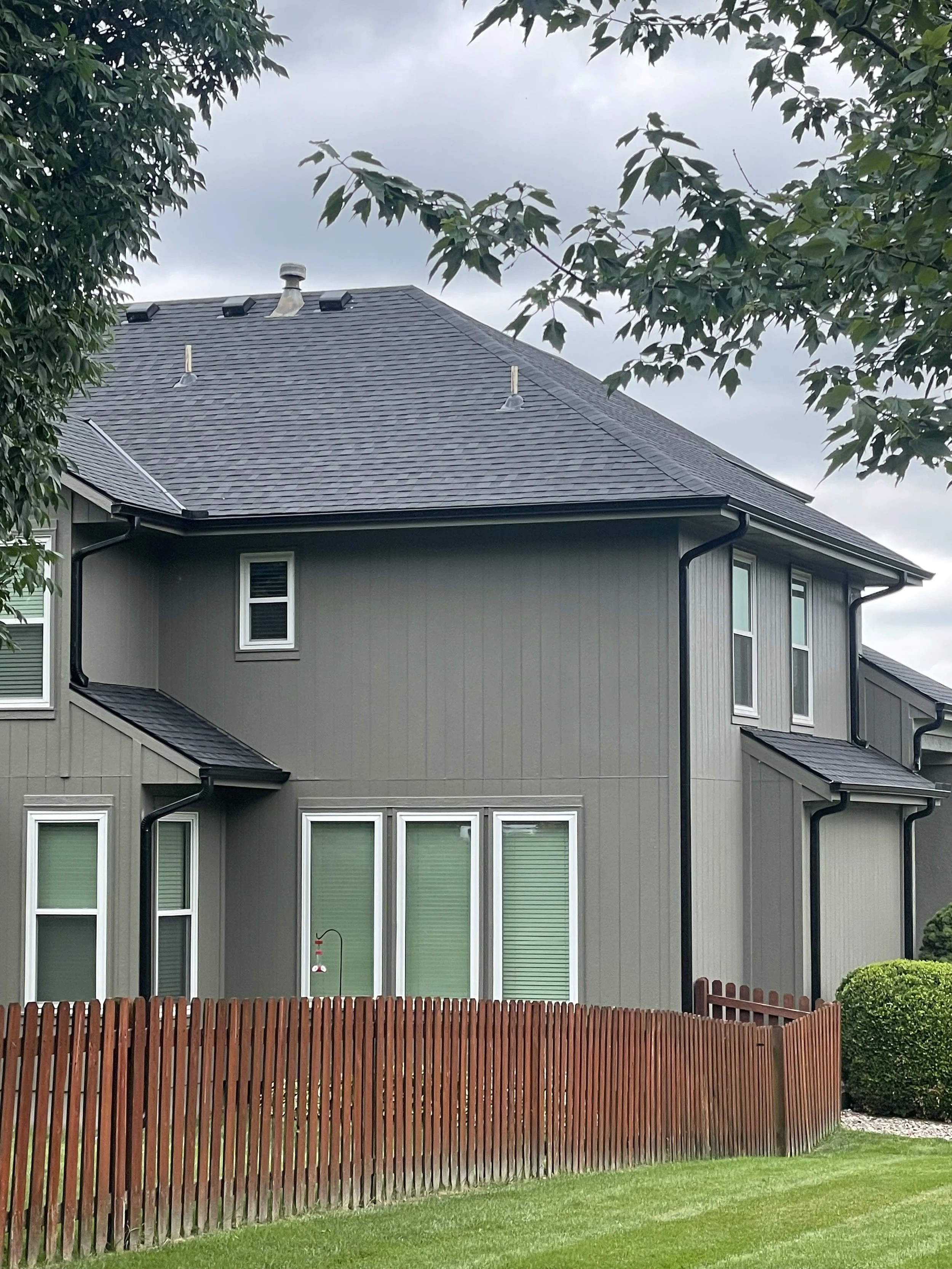 A two-story house with gray exterior walls, black roof, and white window frames, surrounded by a red wooden fence, green grass, and trees.