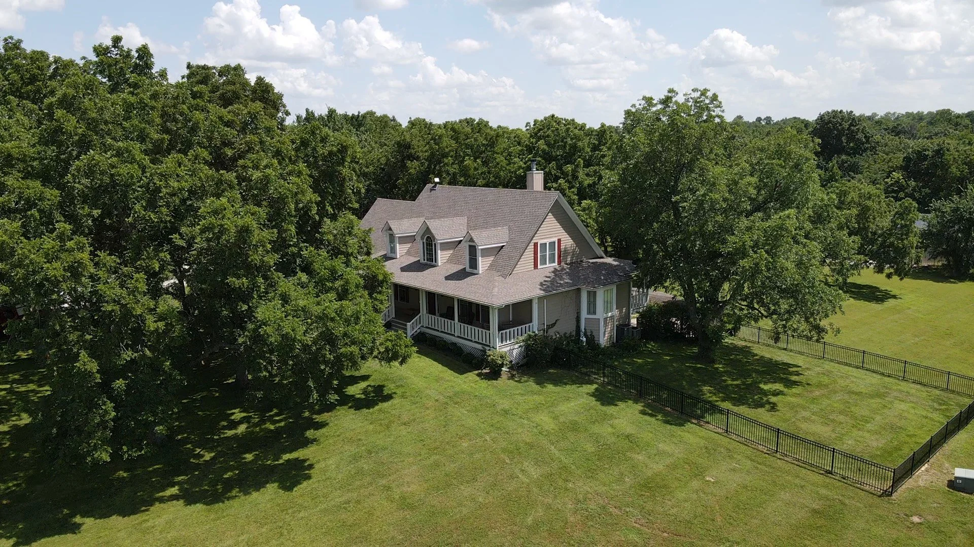 A large house with a gray roof, beige siding, and white porch railing is surrounded by tall green trees and a fenced yard on a sunny day with partly cloudy skies.