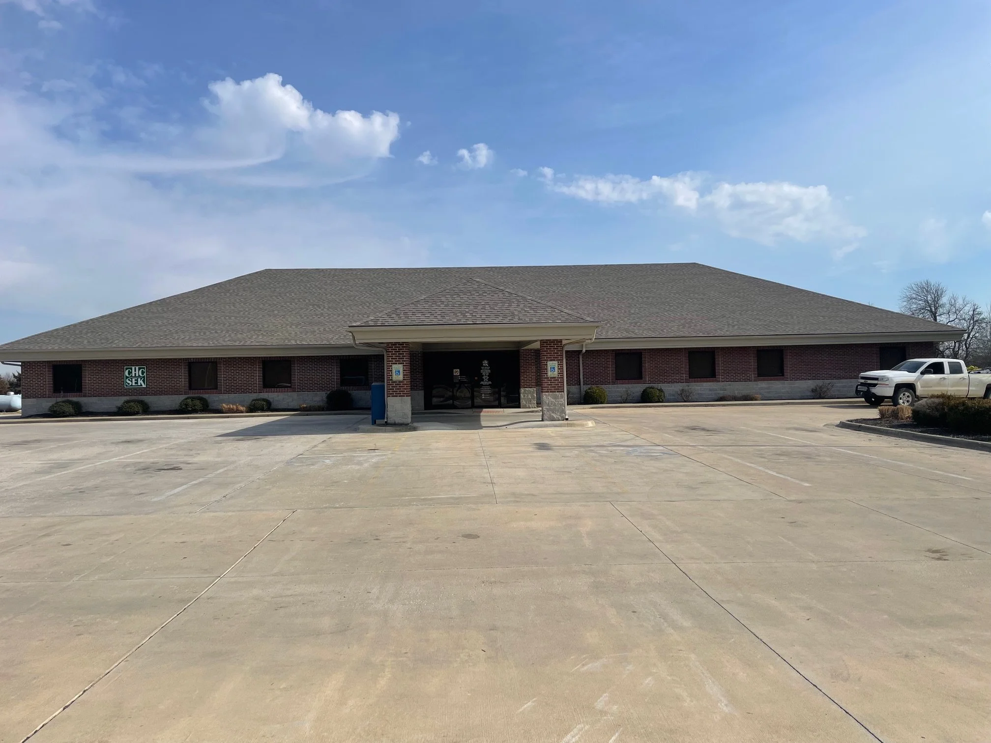 Empty parking lot in front of a single-story brick building with a gabled roof. The building has small bushes along its base and a sign with 'CH' and 'SK' visible on the left. To the right, a white pickup truck is parked. The sky is partly cloudy with blue and white clouds.