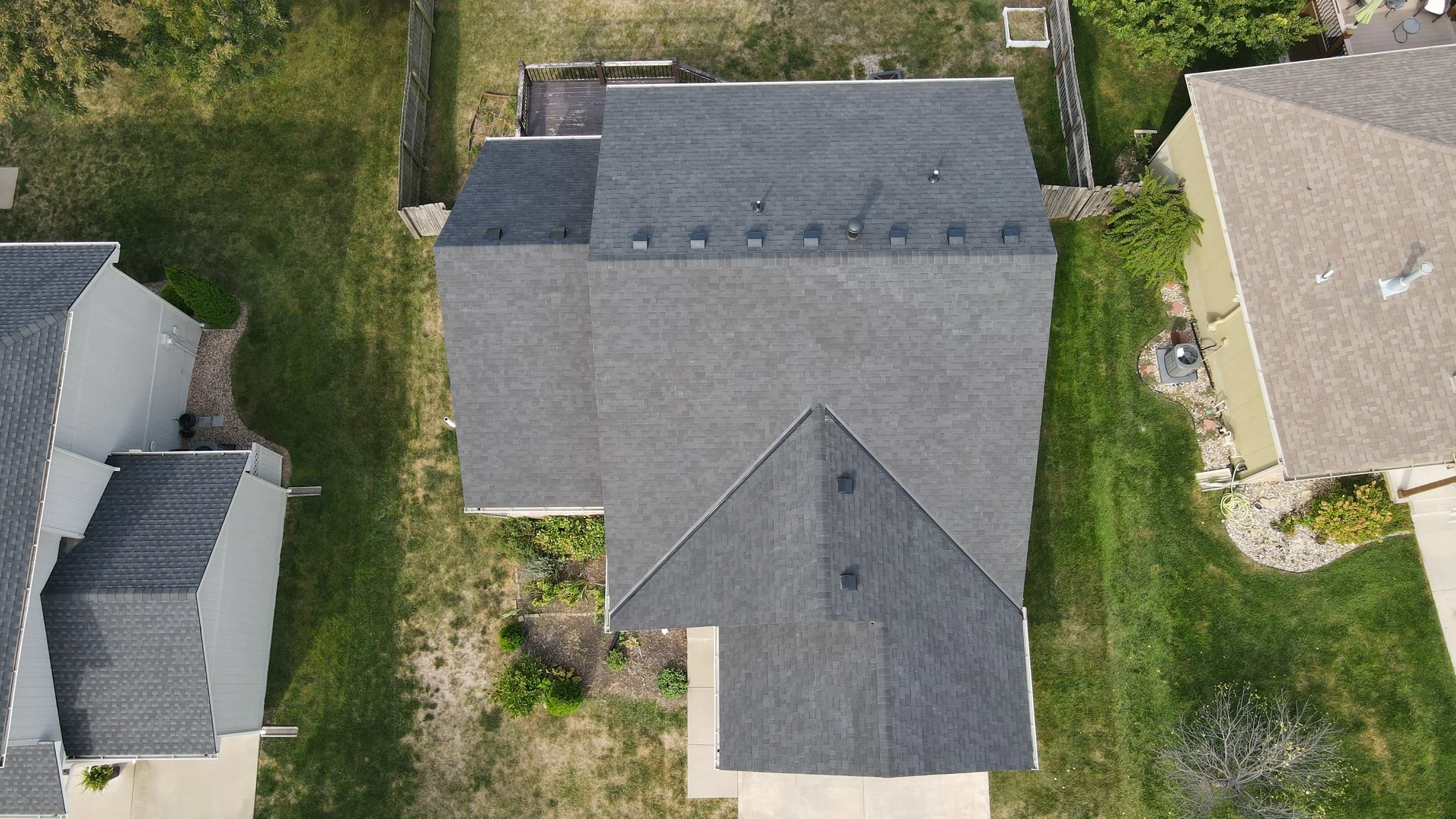An aerial view of a house with a gray roof, surrounded by green grass and neighboring houses with similar roofs in a suburban neighborhood.