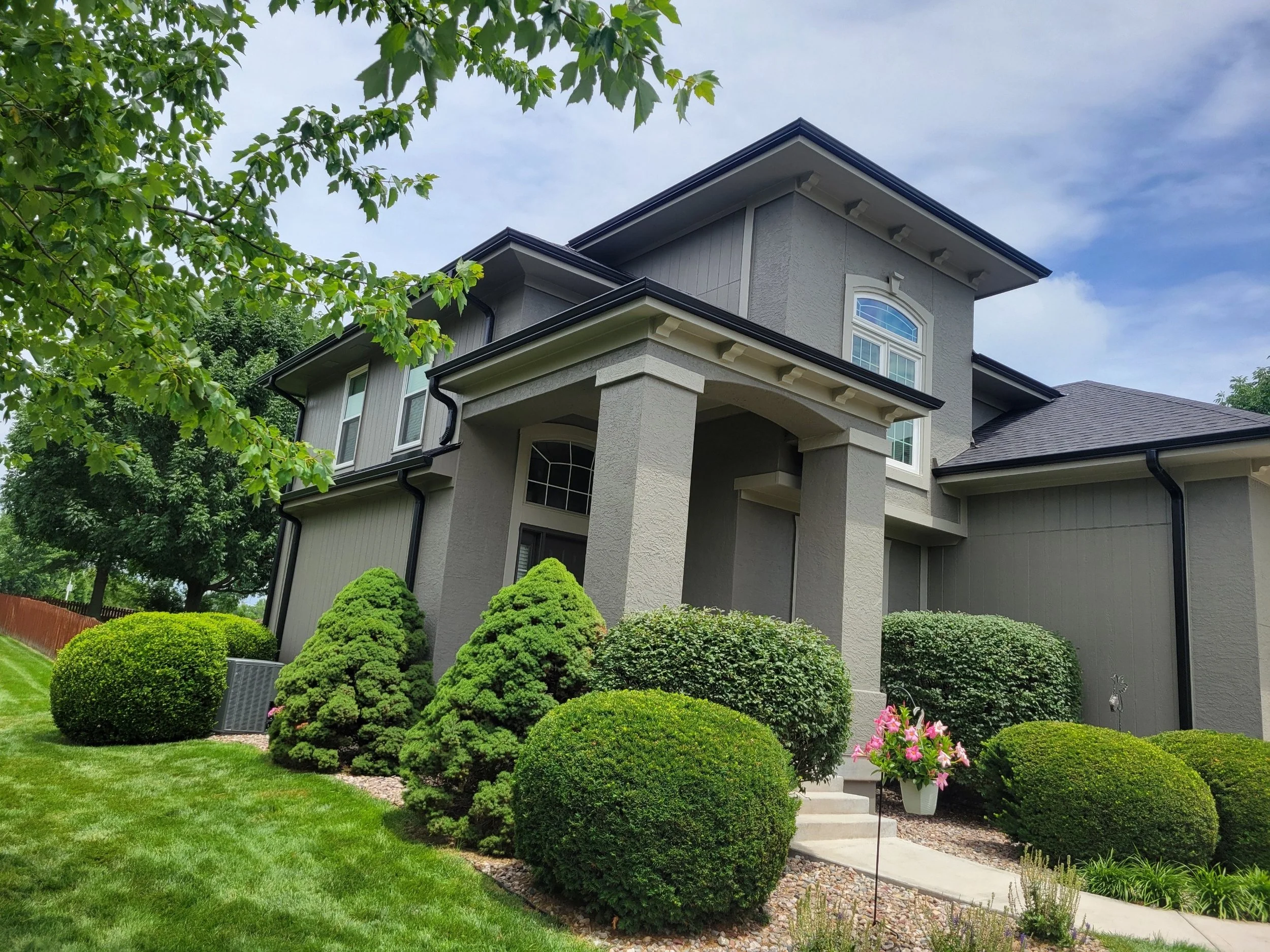 Front view of a modern gray house with well-manicured bushes, green lawn, and a flowerpot with pink flowers.
