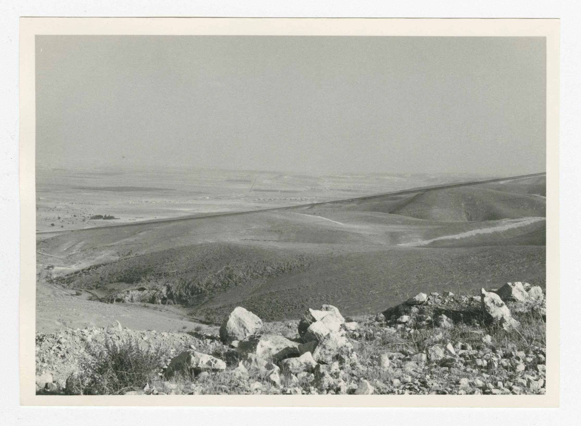 Black and white photo of a vast landscape with rolling hills, rocky terrain in the foreground, and sparse vegetation under an overcast sky.