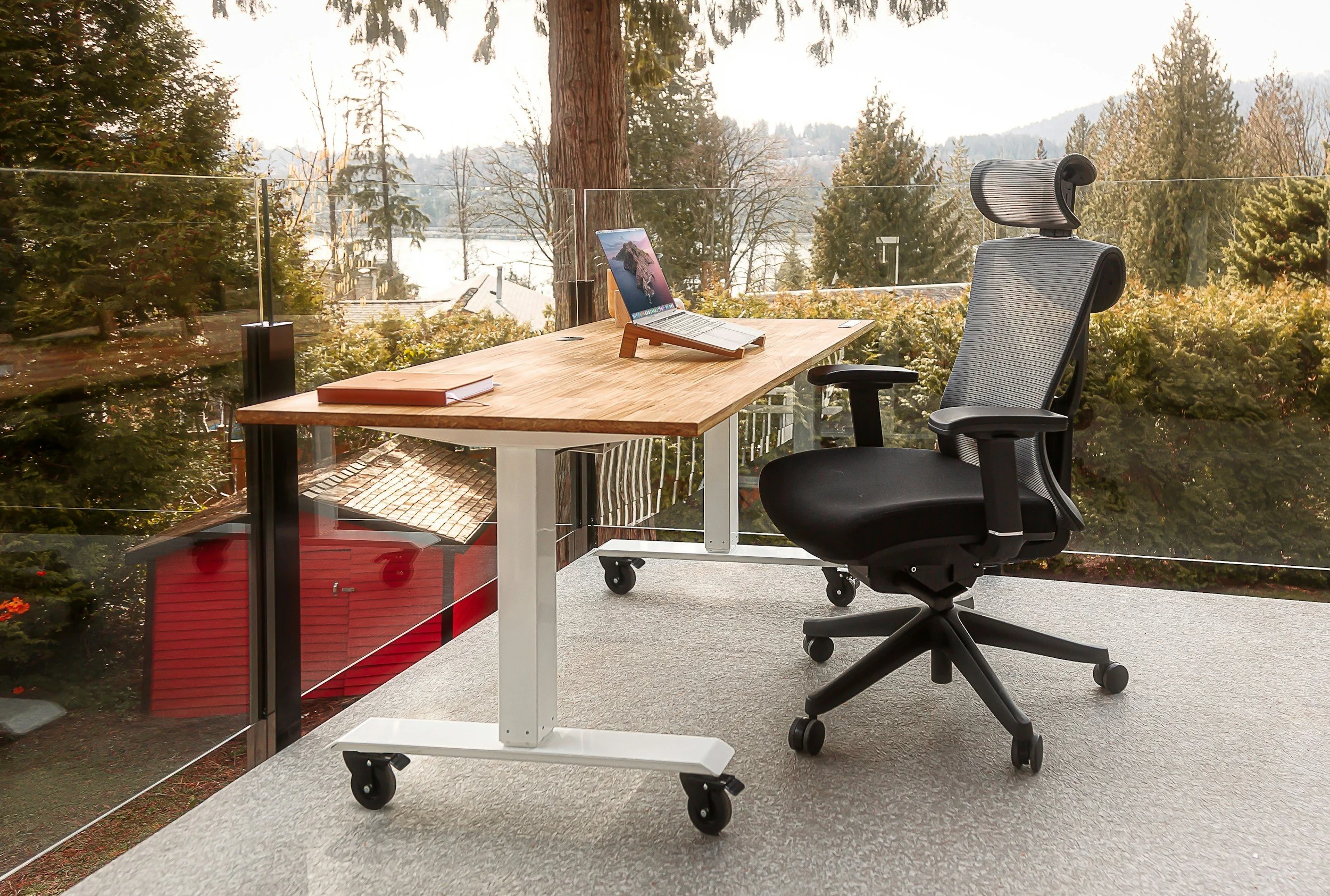 An office desk with a laptop, a closed notebook, and a desk stand, paired with a black ergonomic office chair, set against a glass railing overlooking trees and a winter landscape.