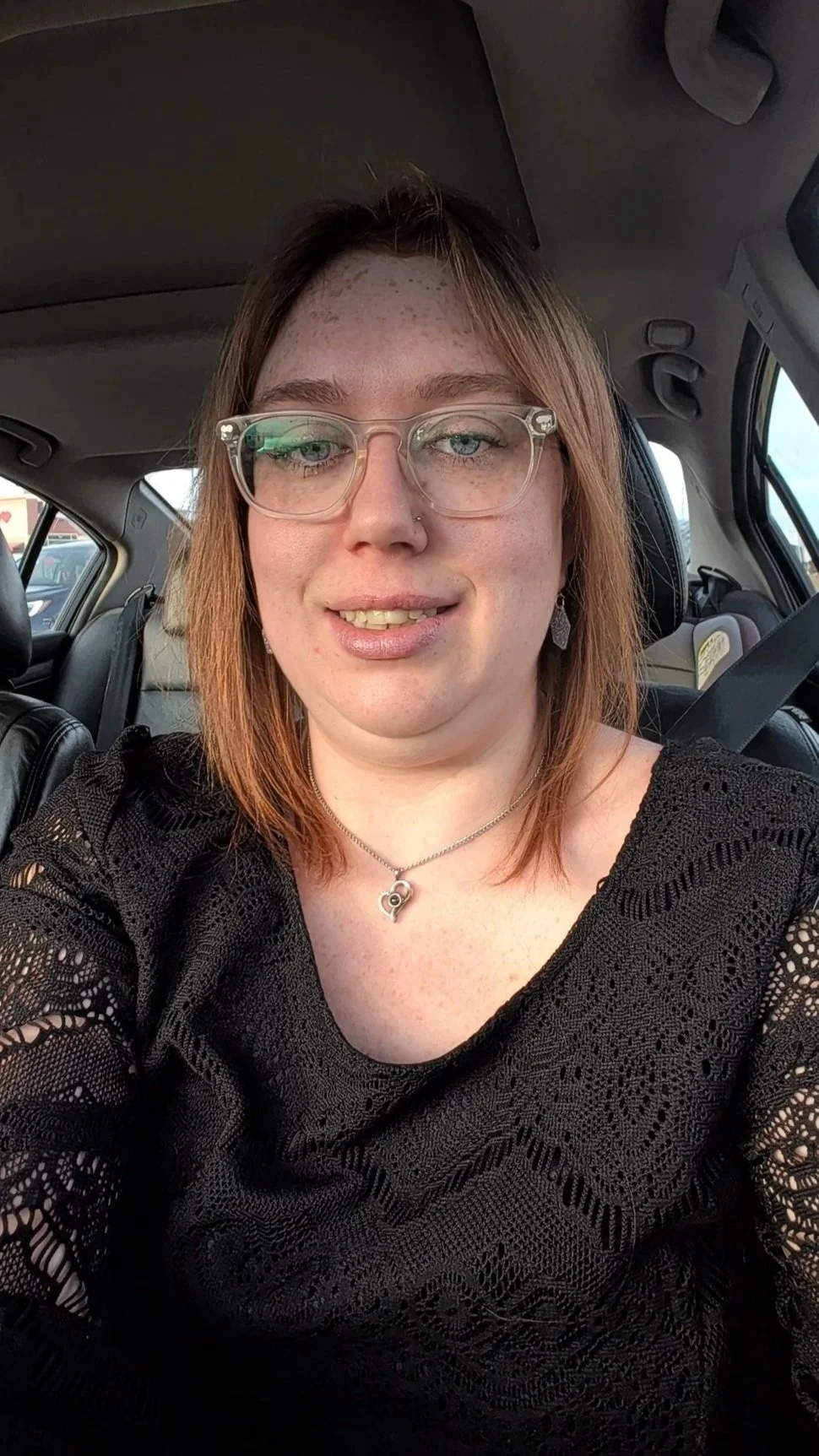 A woman with glasses and earrings sitting in a car, wearing a black lace top and a heart-shaped pendant necklace, smiling at the camera.