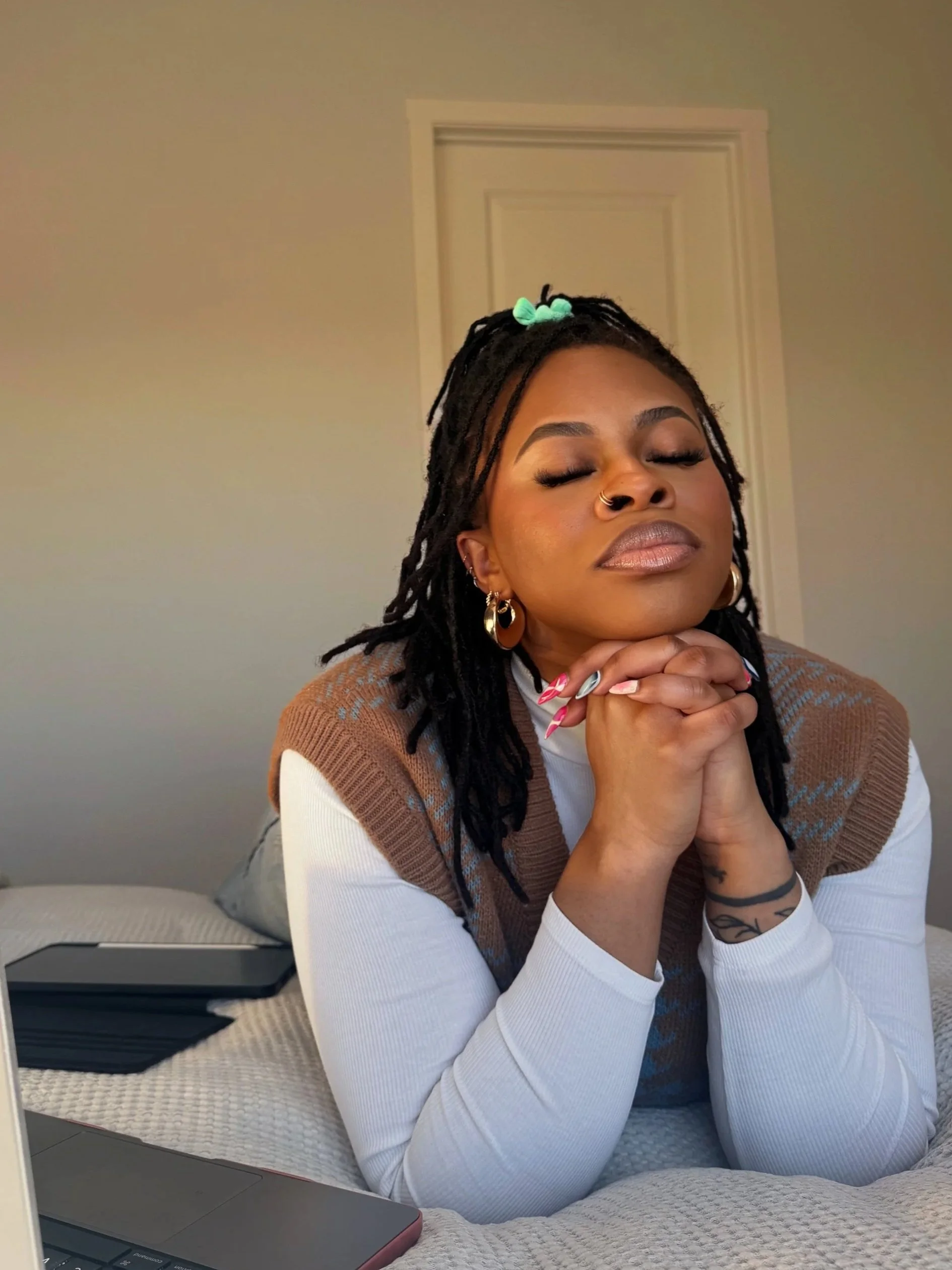 A woman with dreadlocks, wearing a white long-sleeve shirt and a brown sweater vest, has her eyes closed and hands clasped under her chin, sitting on a bed with a laptop nearby.
