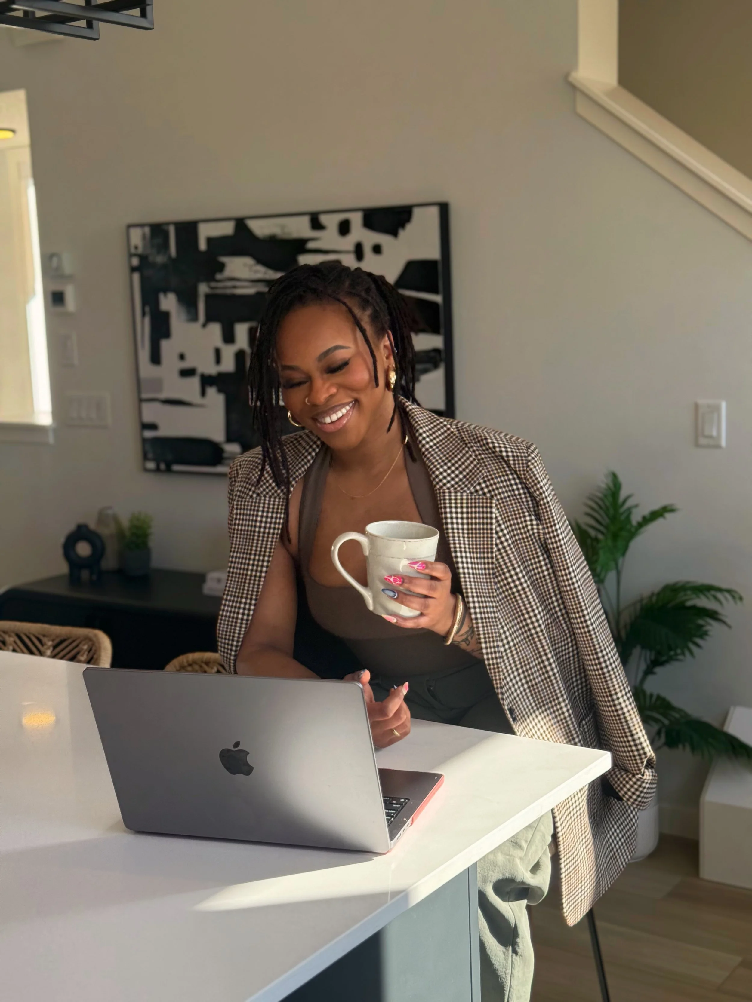 A woman with dreadlocks smiling while holding a mug and looking at a laptop on a white kitchen counter.