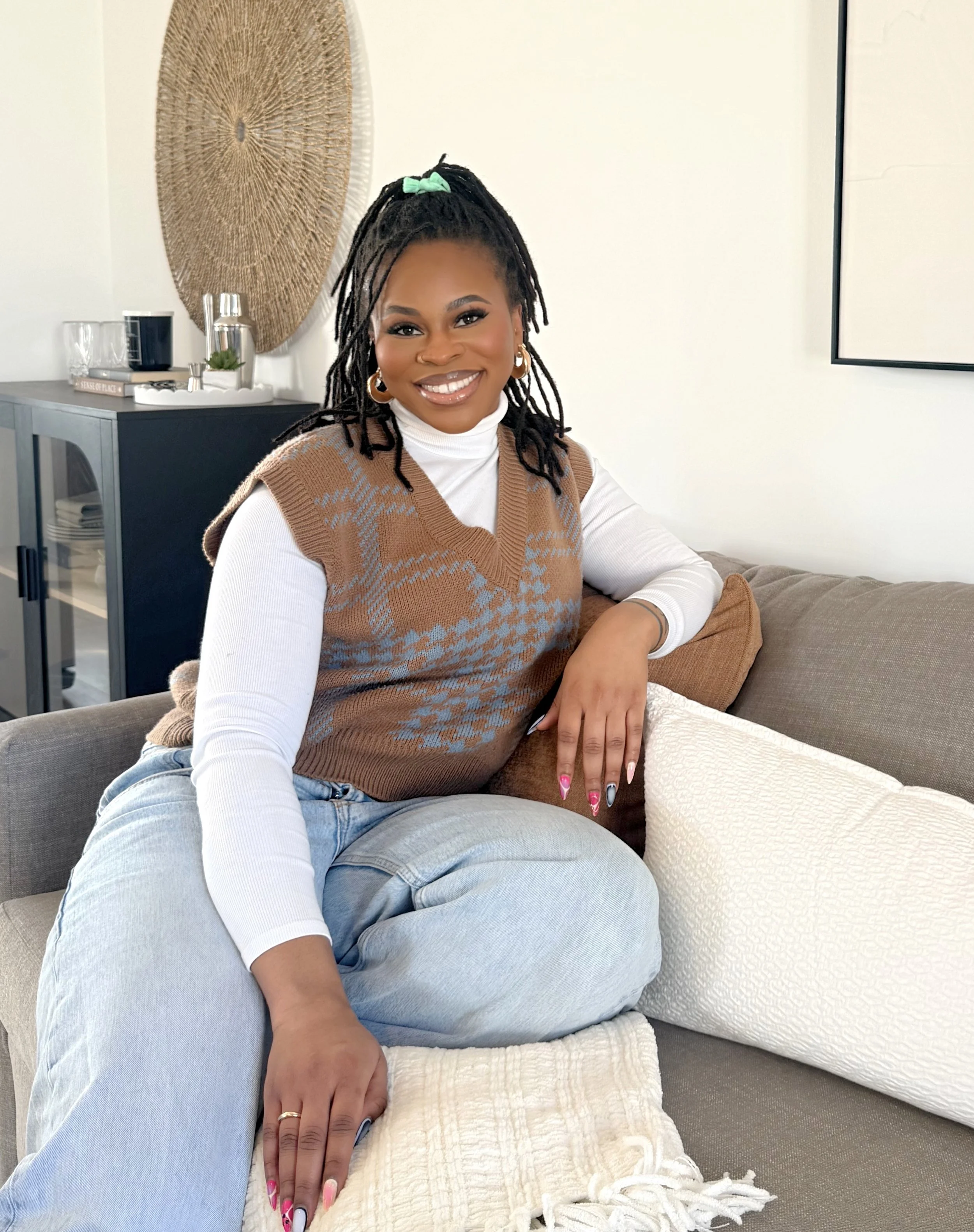A woman with dark skin and dreadlocks styled with a green hairtie, smiling and sitting cross-legged on a gray couch with beige and white pillows. She is wearing a white turtleneck and a brown and blue argyle sweater vest. Behind her is a black cabinet with decorative items, a large woven wall hanging, and part of a white wall with a frame.