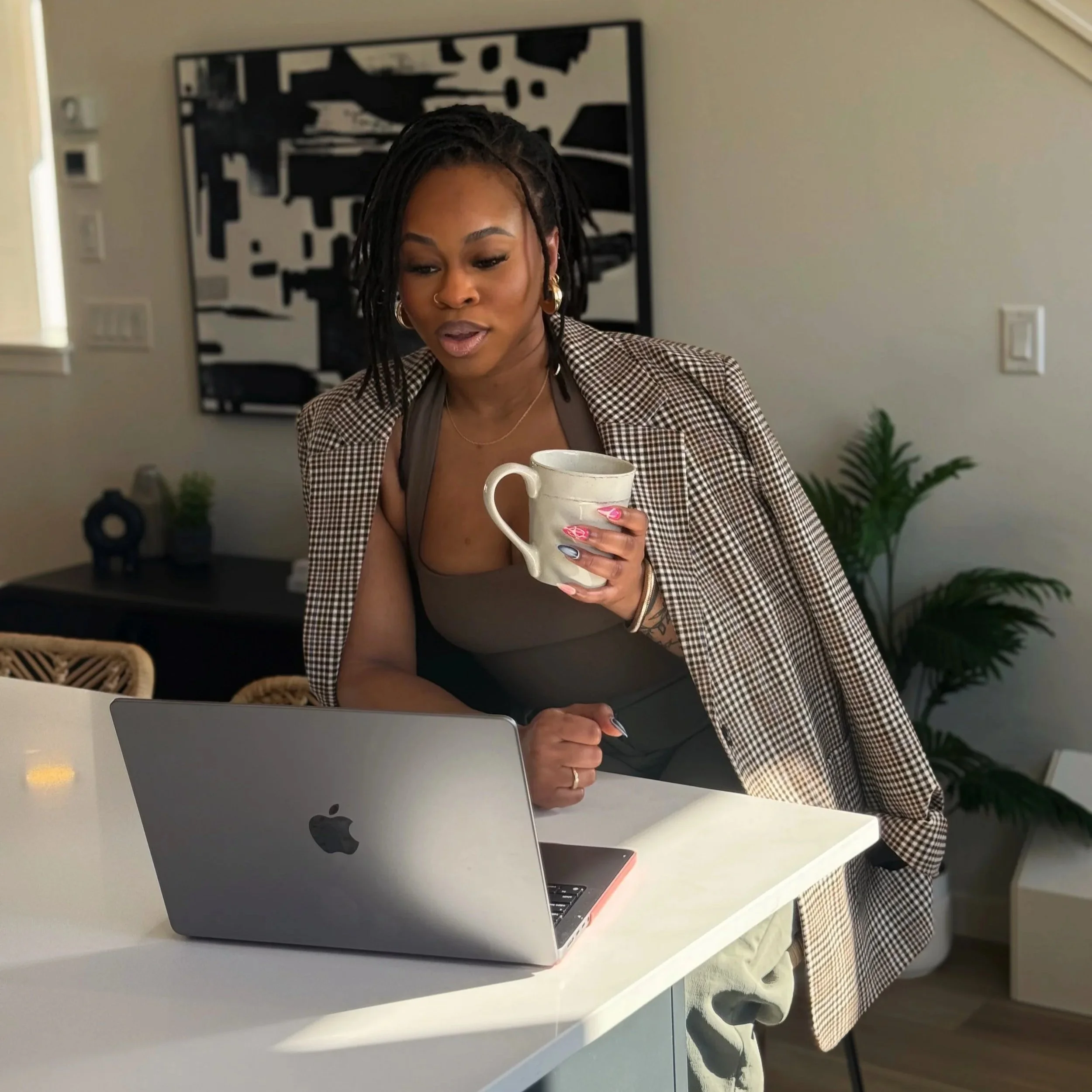 A woman with dreadlocks, wearing a plaid blazer and a brown top, holding a coffee mug while working on a silver MacBook in a modern, well-lit room with neutral decor and a plant in the background.