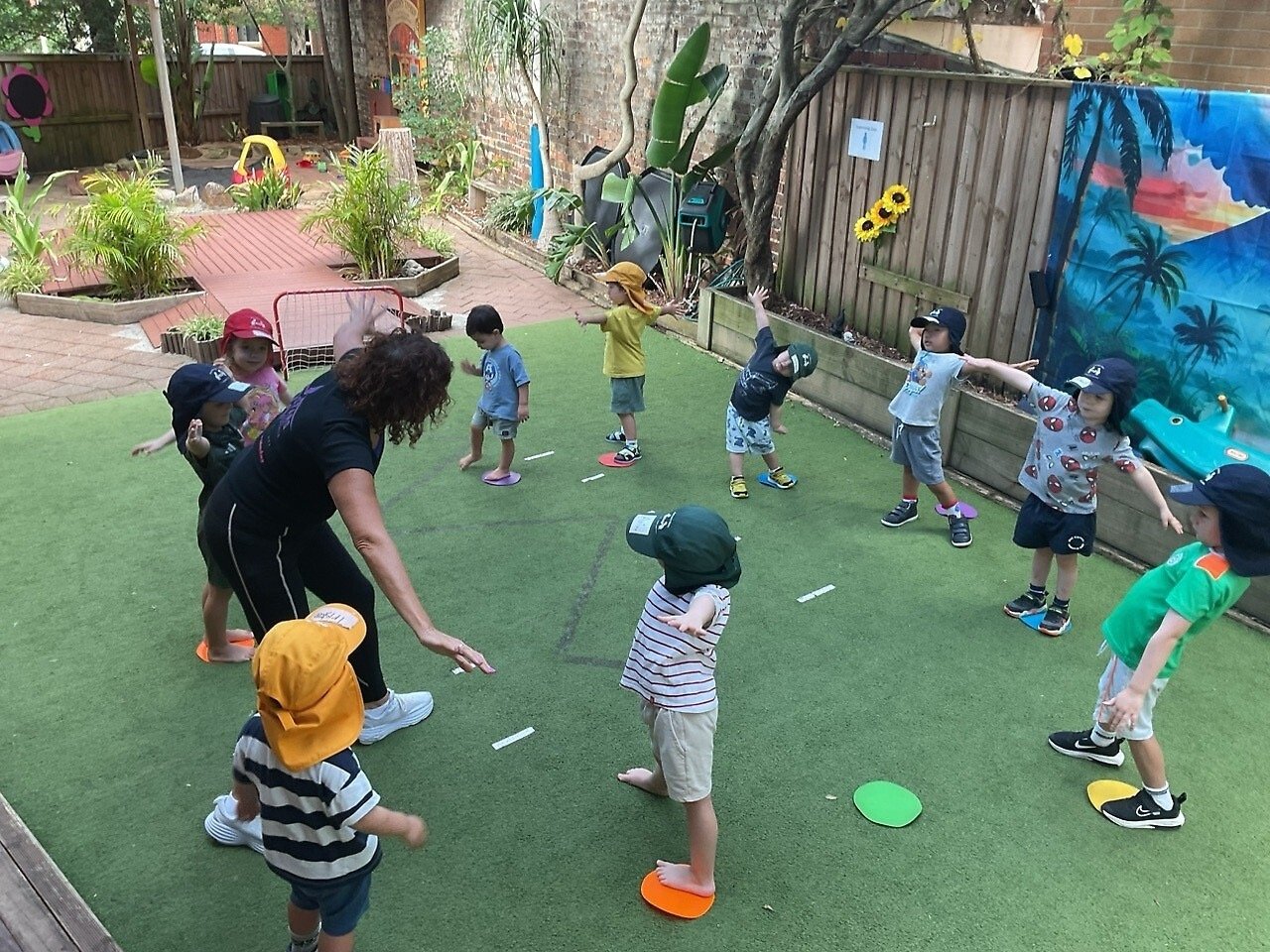 Group of young children and a teacher outdoors on a grassy area, participating in a stretching activity. The children are wearing hats and casual clothes, some standing on colorful plates. The scene is in a play yard with plants, a small goal net, an
