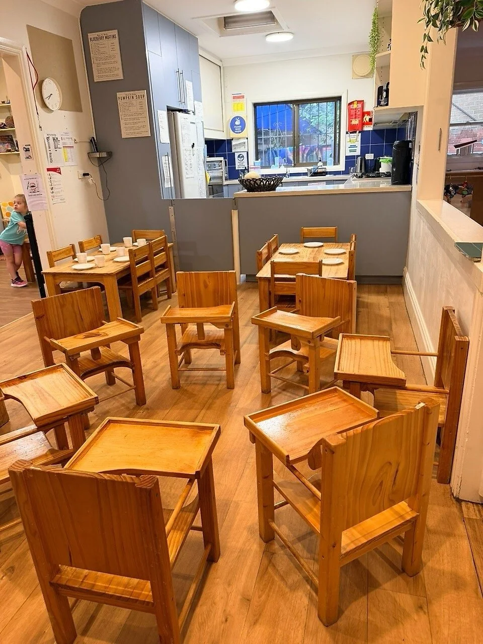 Empty wooden chairs and tables in a cozy cafe or restaurant setting, with a kitchen area in the background featuring a sink, coffee machine, and window.