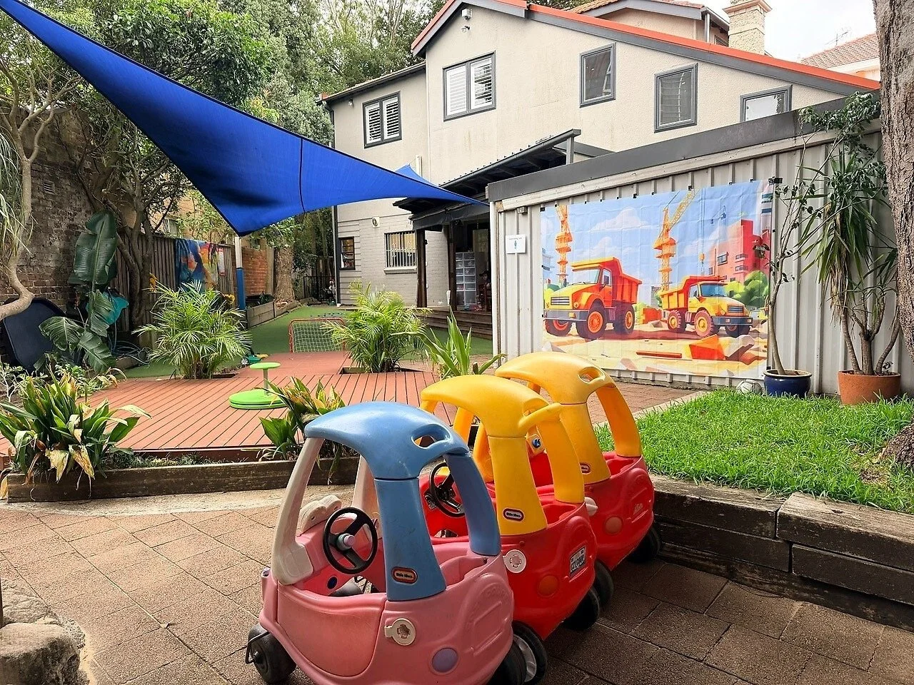 Three children's ride-on toy cars in pink, yellow, and blue parked on a brick patio in a backyard with a wooden deck, plants, and a backdrop featuring a construction-themed blanket with dump trucks.