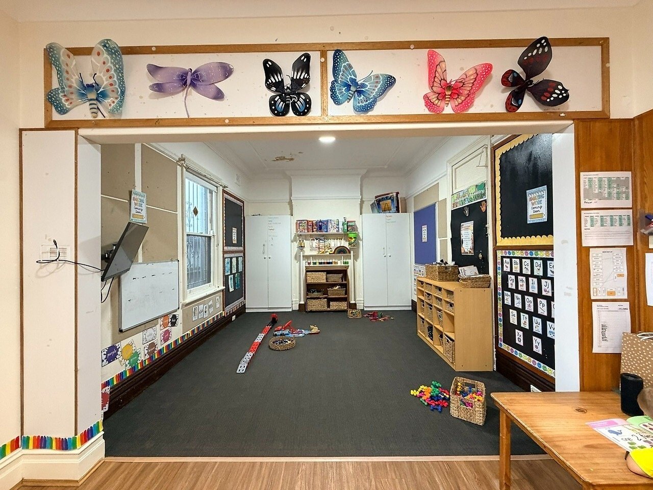 Classroom with butterfly decorations on the doorway, blackboard, shelves with toys, and scattered building blocks on the carpeted floor.