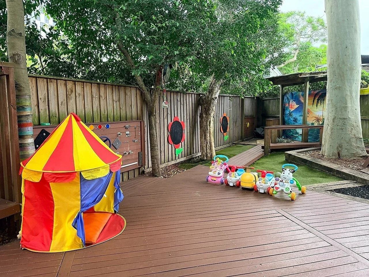Children's outdoor play area with a yellow and red circus tent, a line of toy ride-on animals, and colorful wall decorations featuring flowers and tropical scenes.