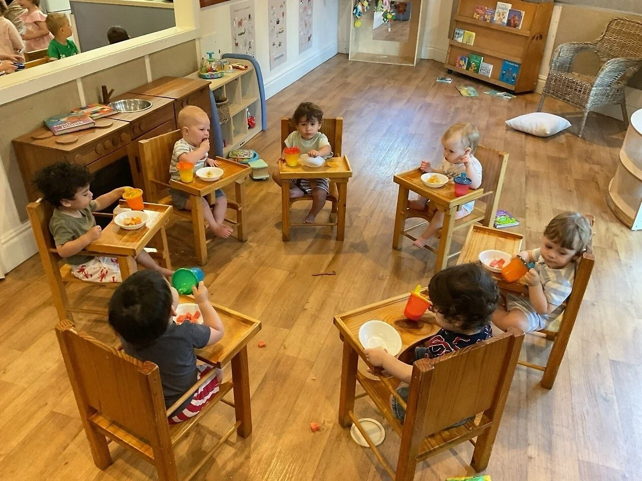 Seven young children sitting in wooden high chairs around a wooden table, eating snacks, in a daycare or preschool setting.