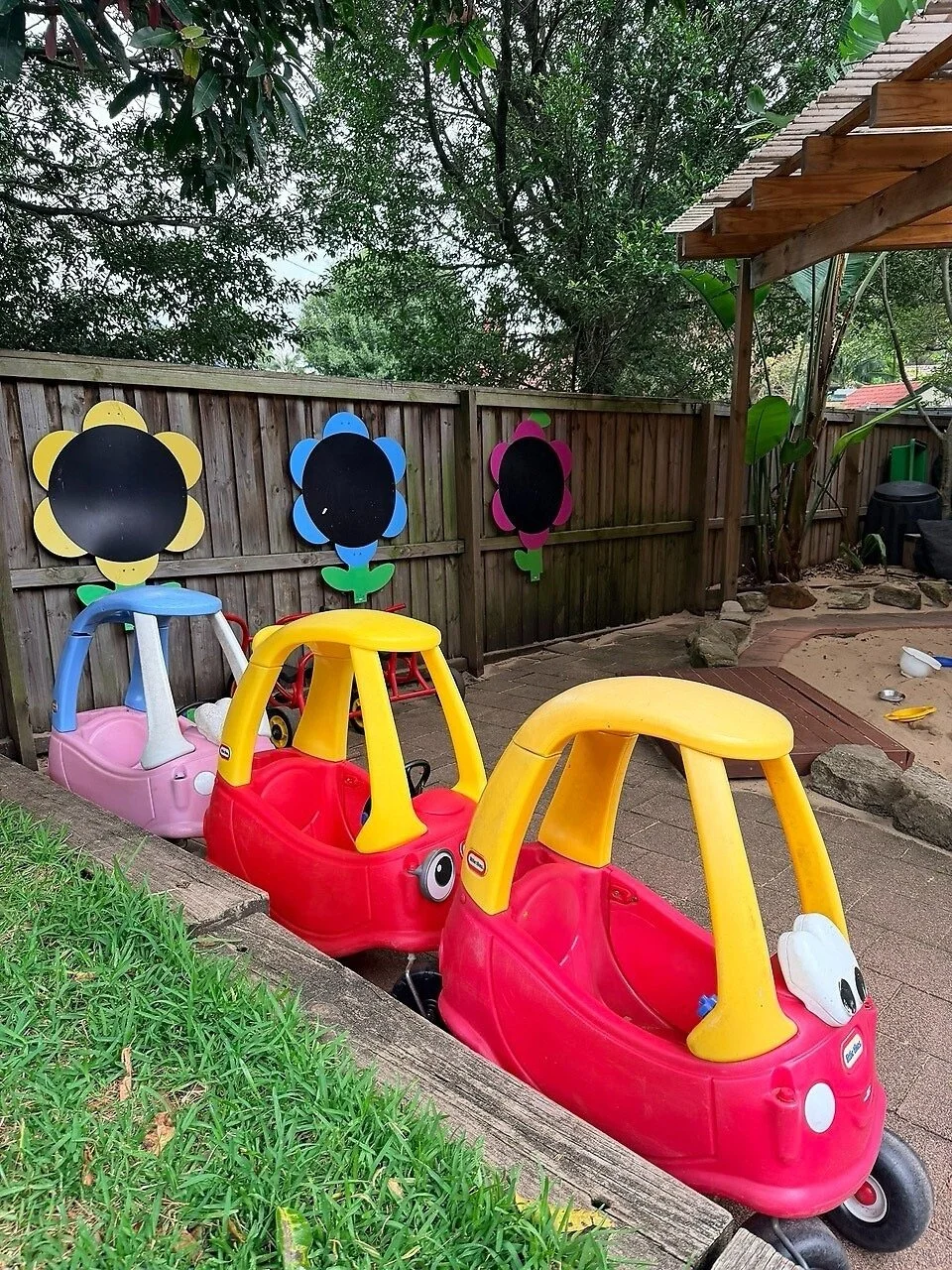 Colorful children's pedal cars parked next to a wooden fence decorated with flower cutouts, in an outdoor play area with a brick pathway and lush trees in the background.