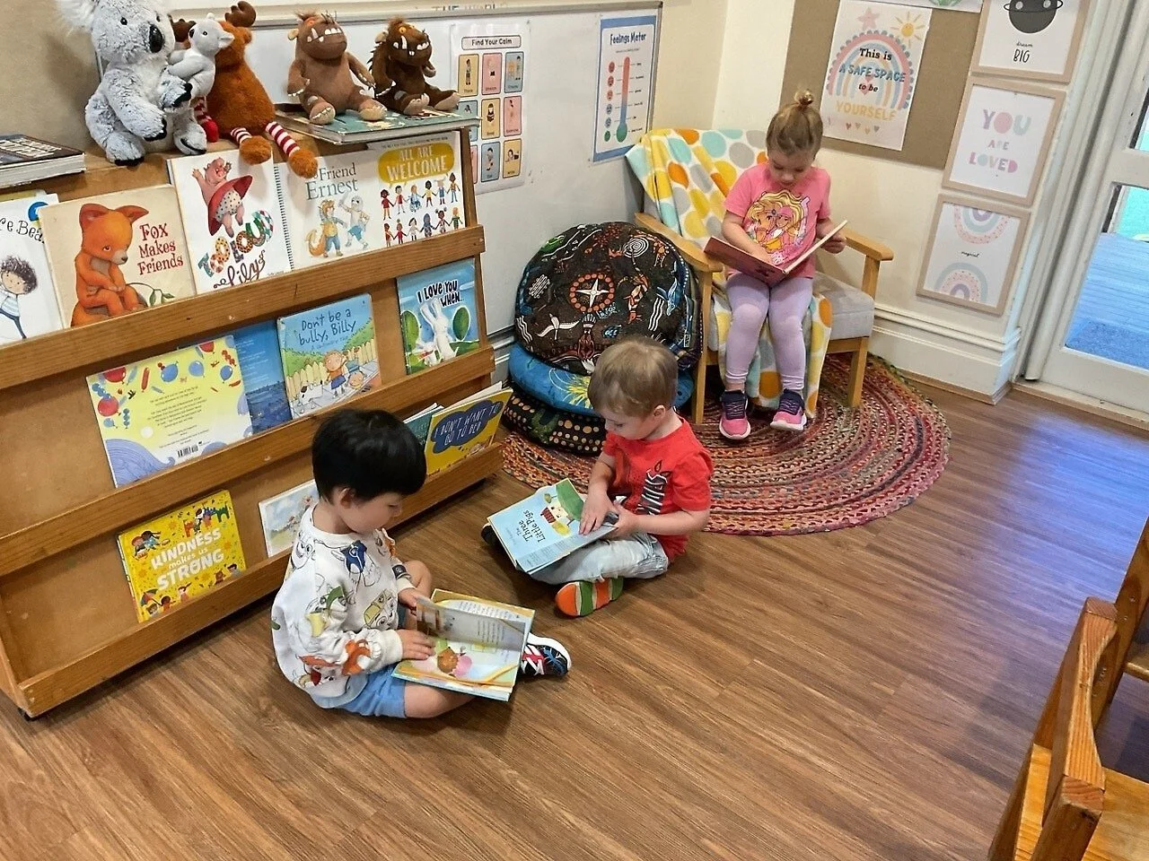 Three children reading books in a cozy corner of a library or classroom, with a bookshelf filled with children's books and plush toys on top, a colorful rug, and framed artwork on the walls.