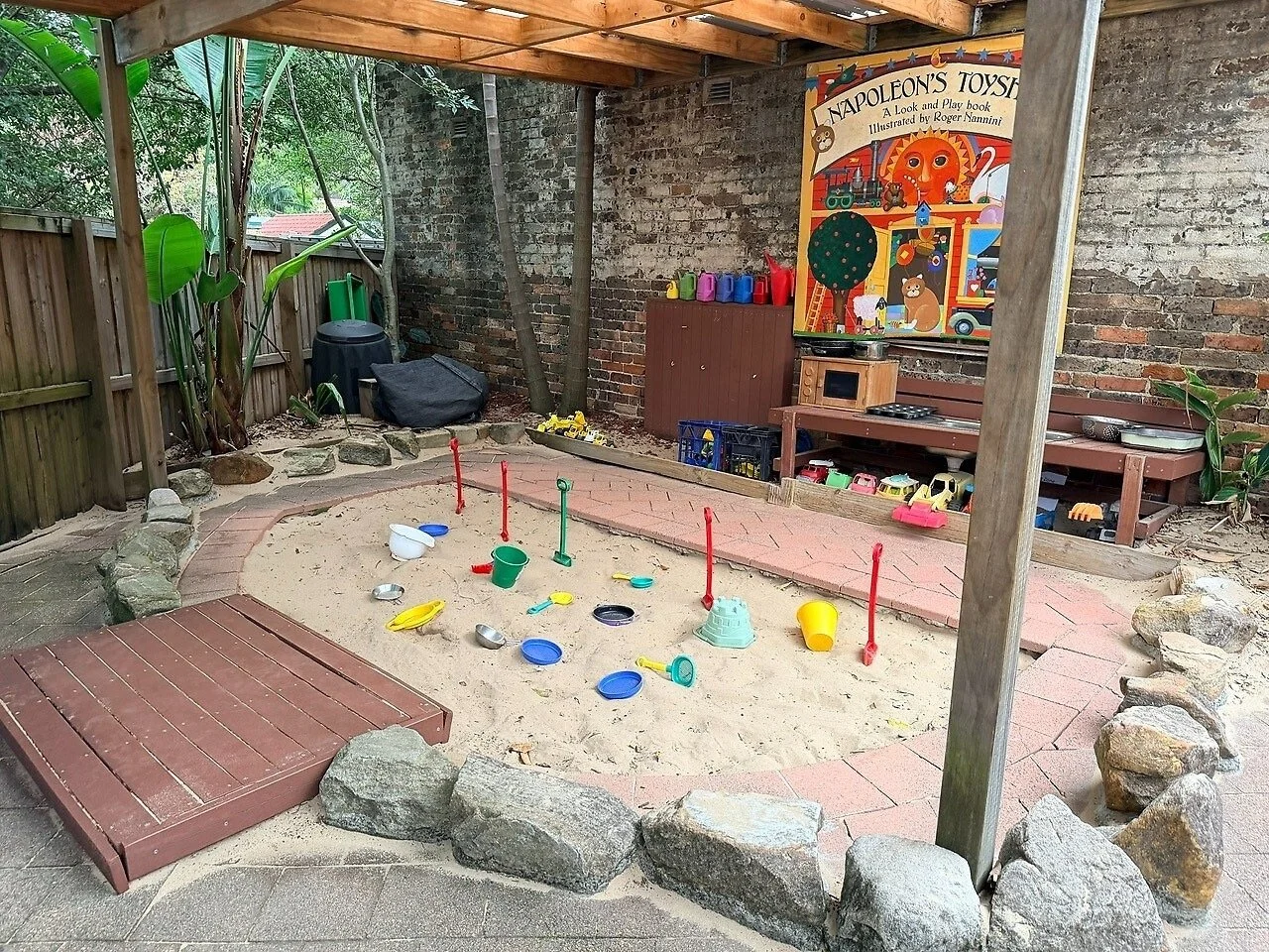 Children's outdoor play area with sandbox, toys, and a wooden playhouse under a shaded structure. A colorful poster art called 'Napoleon's Toy Store' is on the brick wall in the background.