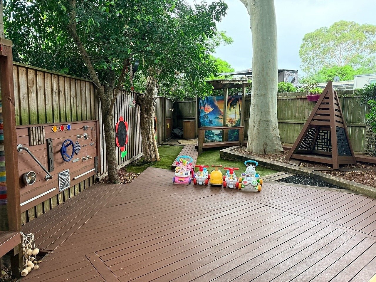 Child's outdoor play area with toys, trees, a wooden deck, and a fence decorated with colorful elements and a tropical mural.