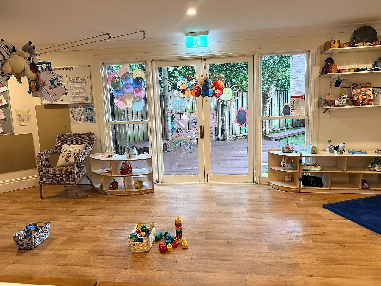 Children's playroom with wooden floors, toys, shelves, drawings, and decorations. Outside is a deck and backyard with trees.