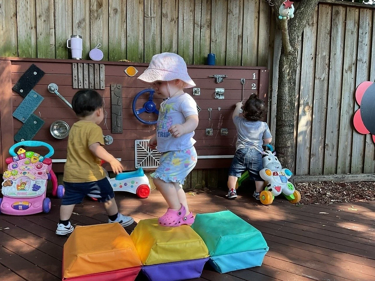 Three young children playing outside on a wooden deck with toys and a musical activity wall, surrounded by a wood fence and trees.