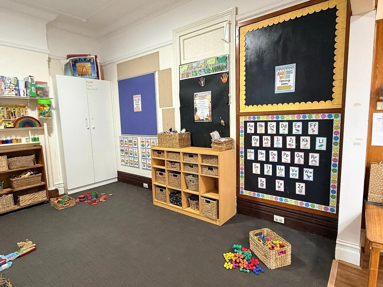 A classroom corner with storage shelves filled with educational toys and materials, a black bulletin board with the alphabet, and various small toys on the carpeted floor.