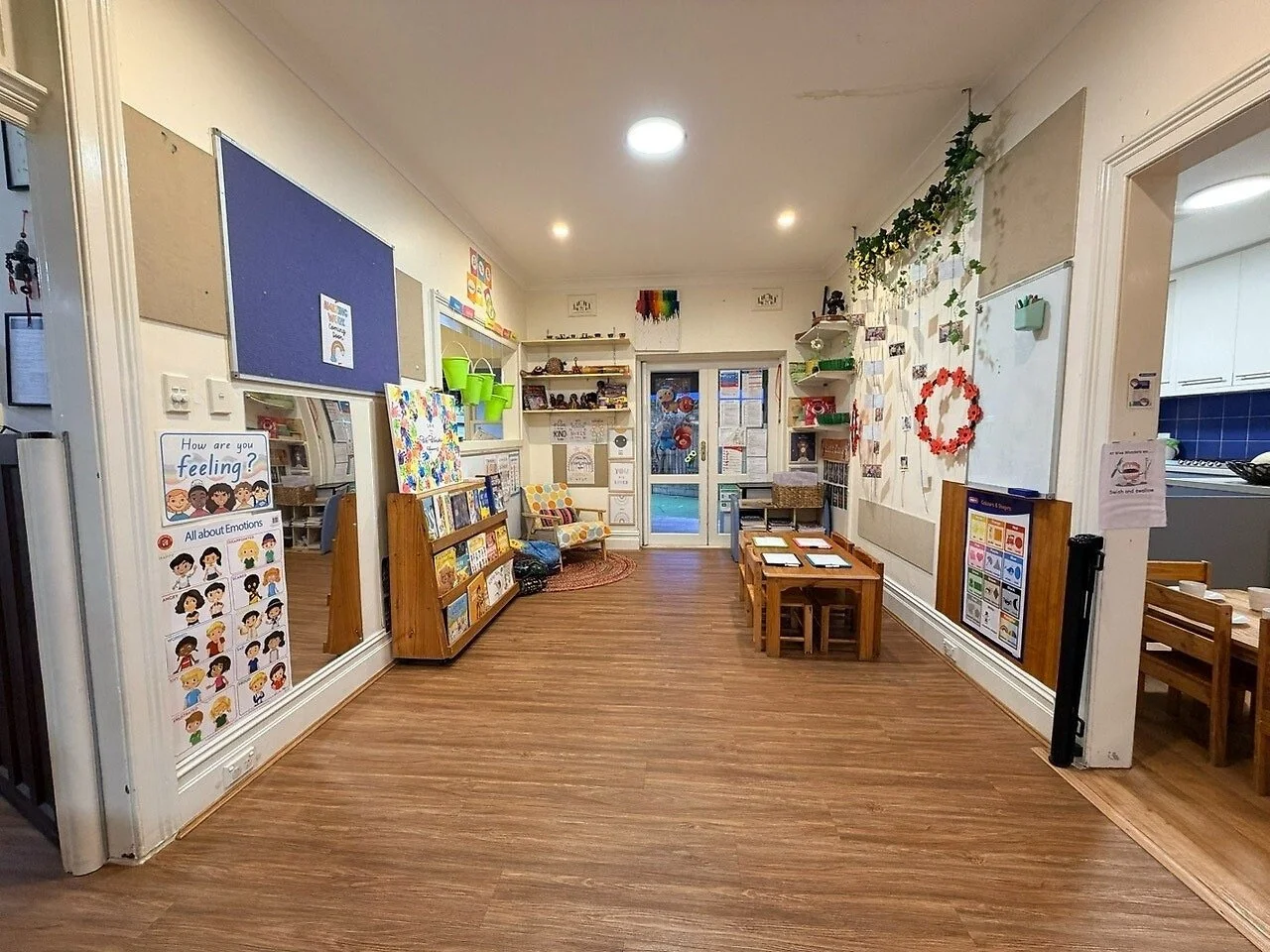 A preschool classroom with wooden floors, a bookshelf filled with children's books, small tables and chairs, colorful decorations, and artwork on the walls.