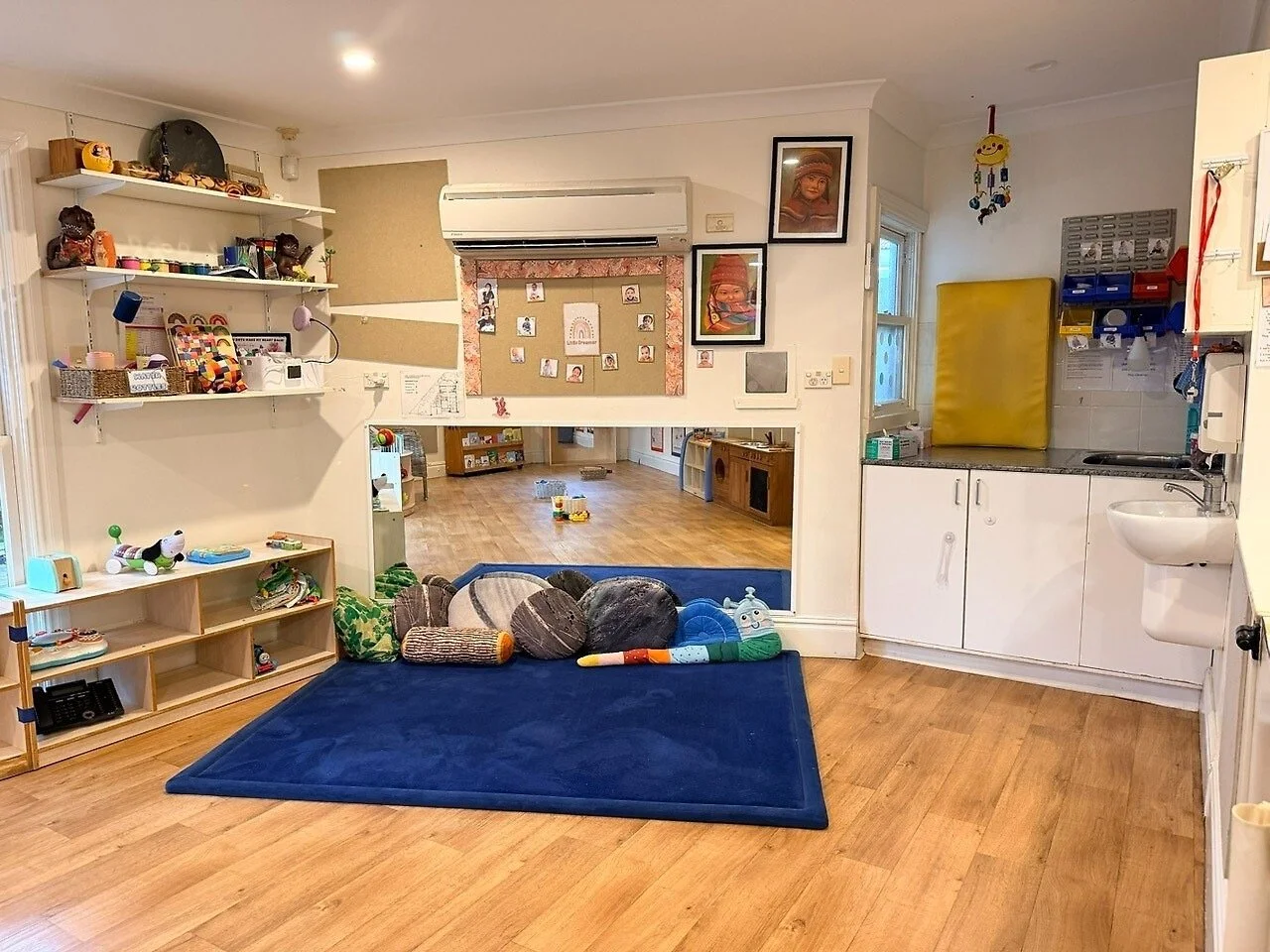 A cozy children's playroom with a blue rug on wooden floors, stuffed toys and pillows, shelves filled with toys and books, framed photographs on the wall, a kitchenette with storage and a yellow padded mat, and various decorations.