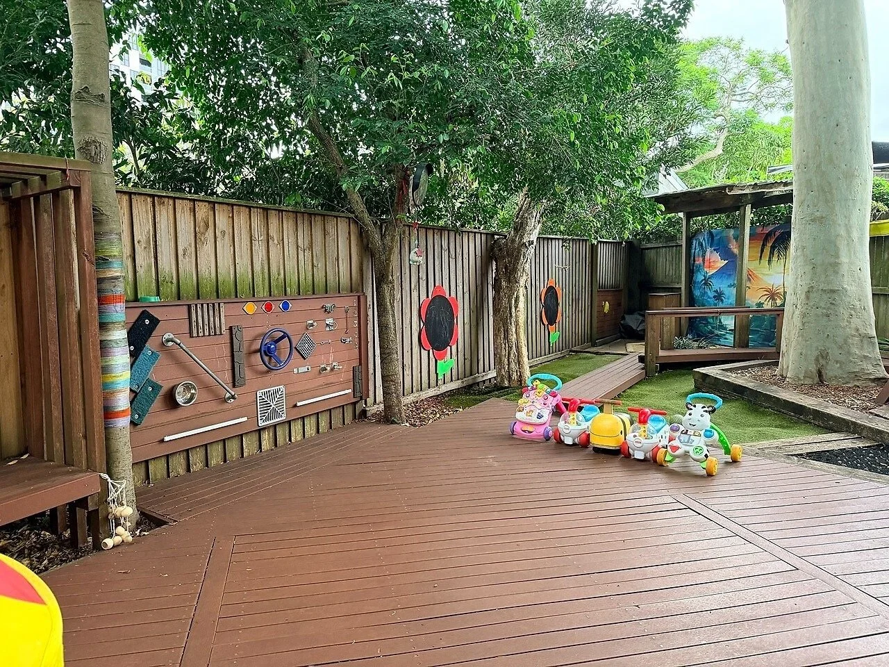 Children's outdoor play area with toy ride-ons, a wooden deck, decorative painted flowers on the fence, trees, and a colorful mural in the background.