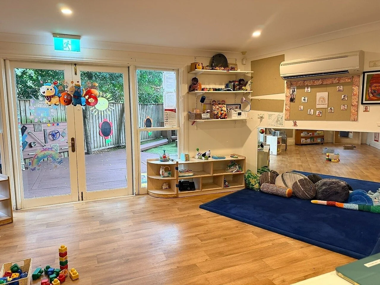 A bright, colorful playroom with wooden flooring and large glass doors leading to an outdoor deck with trees. The room has shelves with toys and books, a blue cushioned area with stuffed pillows, and toys scattered on the floor.