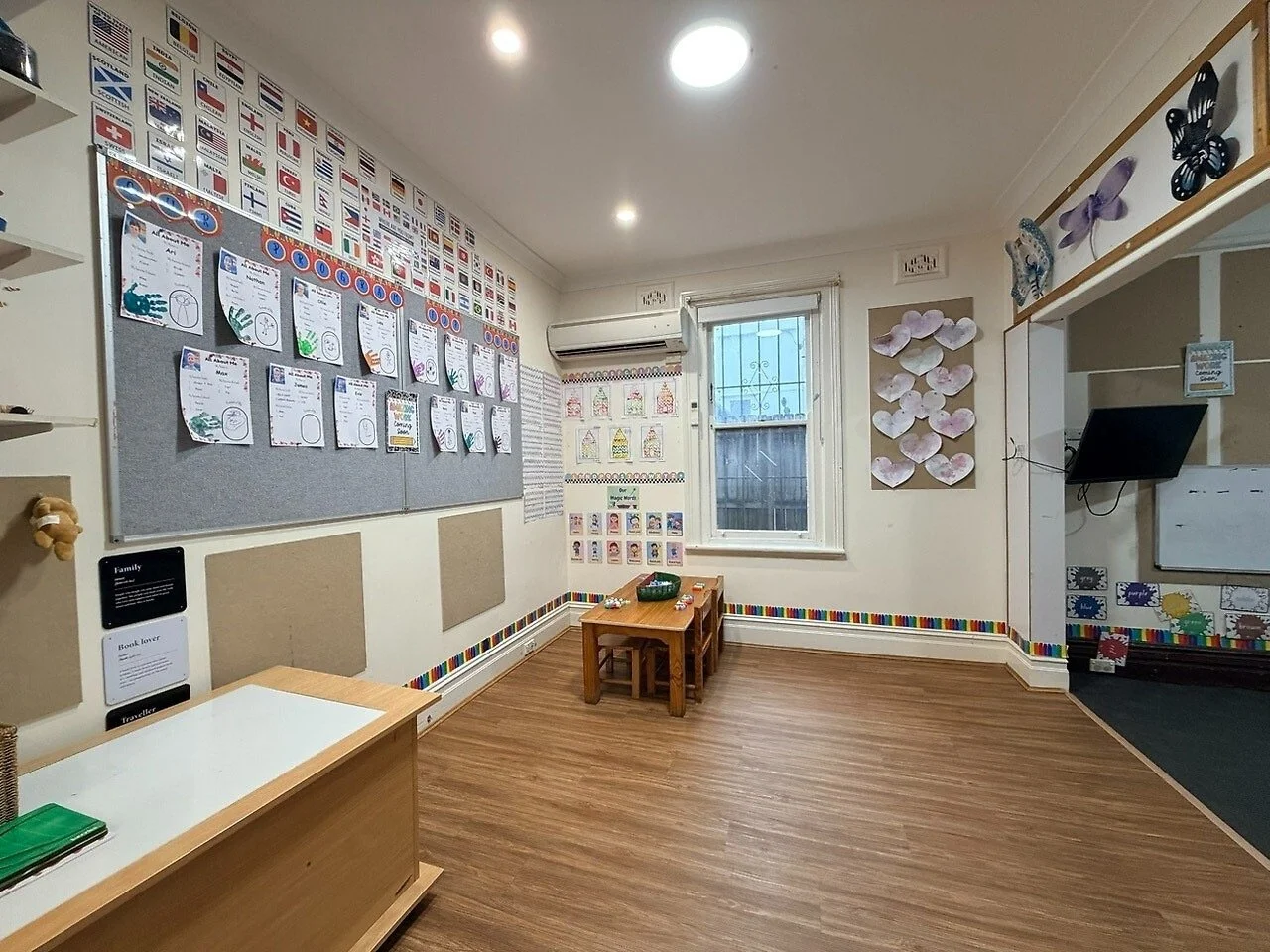Empty classroom with educational decorations, flags, and artwork on the walls, wooden floor, and a small table with chairs.