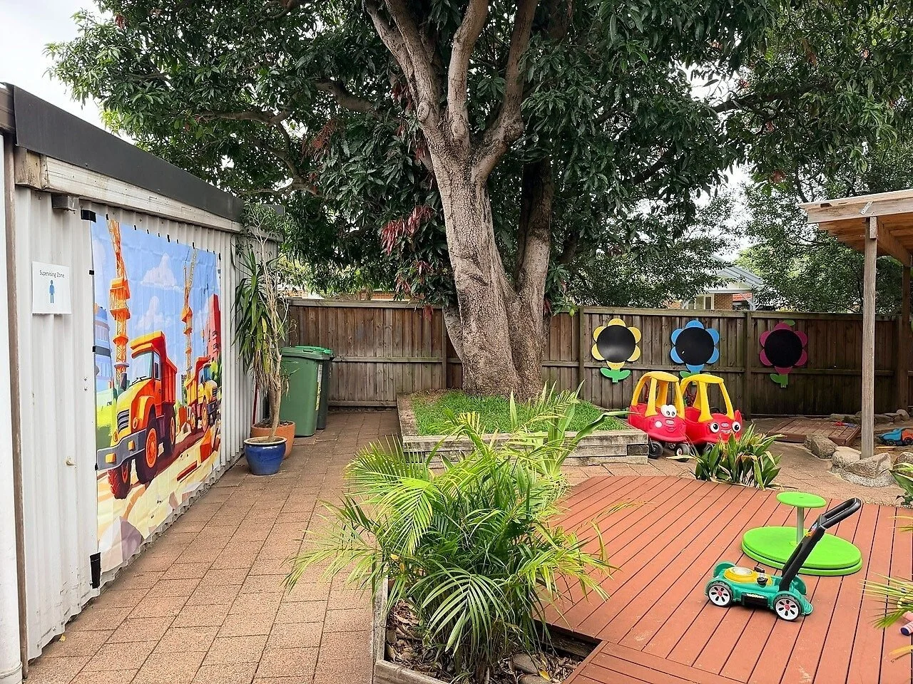 A backyard with a large tree, potted plants, a wooden deck, small toy cars, and a colorful fencing mural of construction trucks.