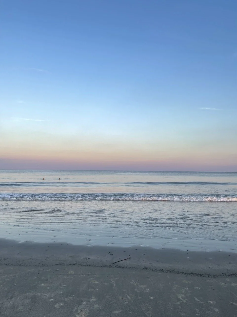 Calm ocean shoreline at sunset with soft pastel sky and gentle waves, two small distant swimmers in the water.