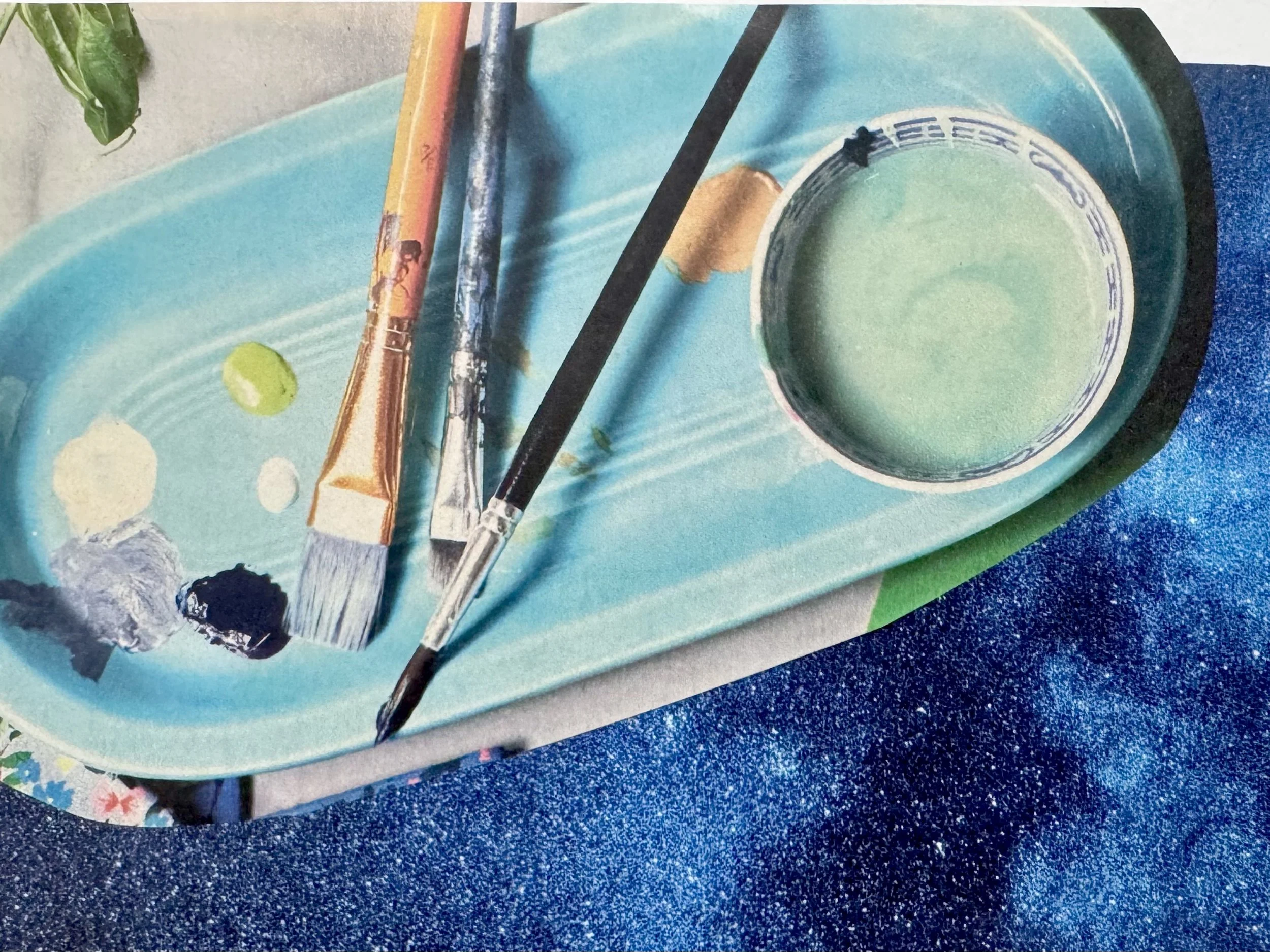 A blue tray with two paintbrushes, a small container of light green paint, black, white, and gray paint blobs, and a small green plant on the side.