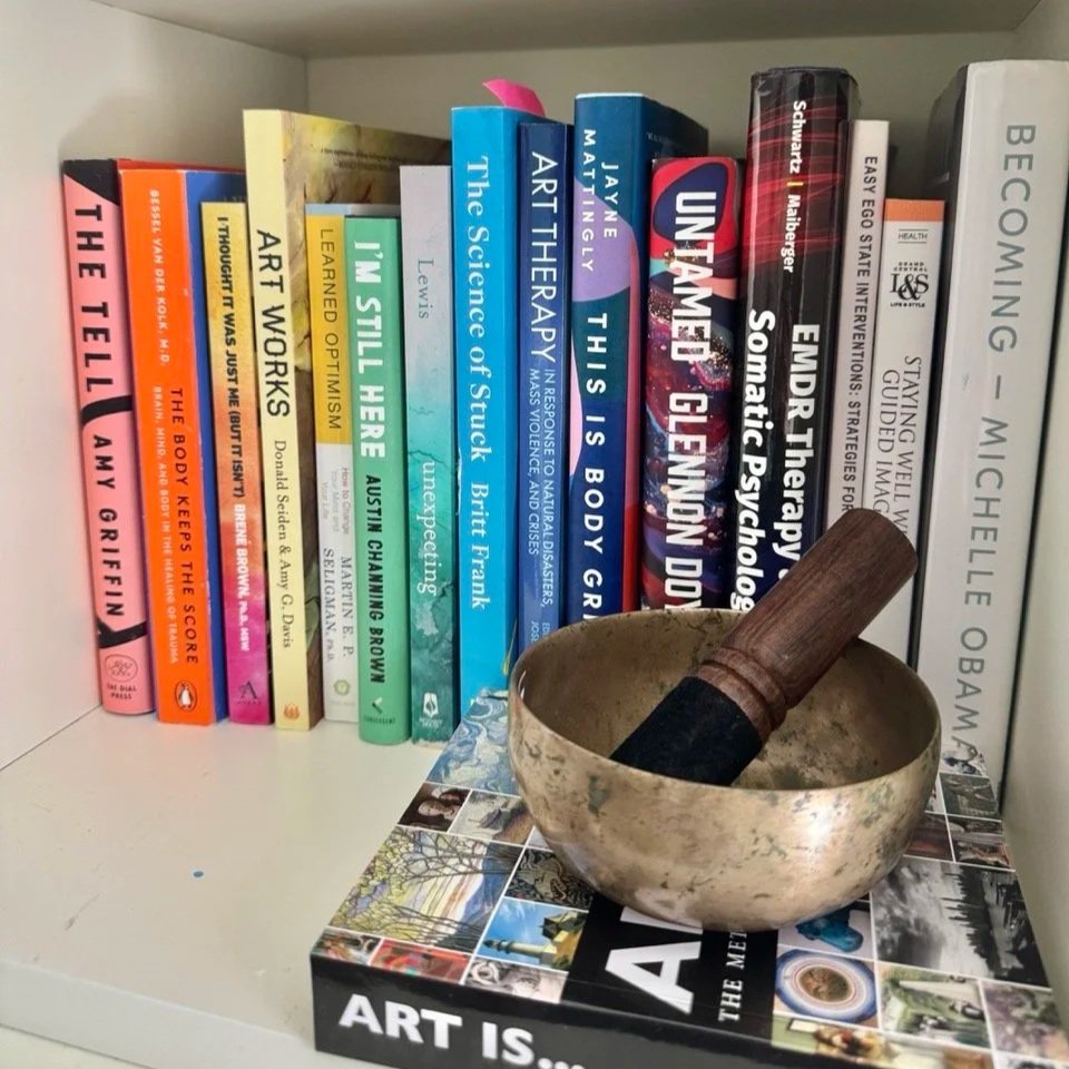 A bookshelf with colorful books on art, psychology, therapy, and health, with a singing bowl and a wooden mallet placed on a book titled 'Art Is...'.