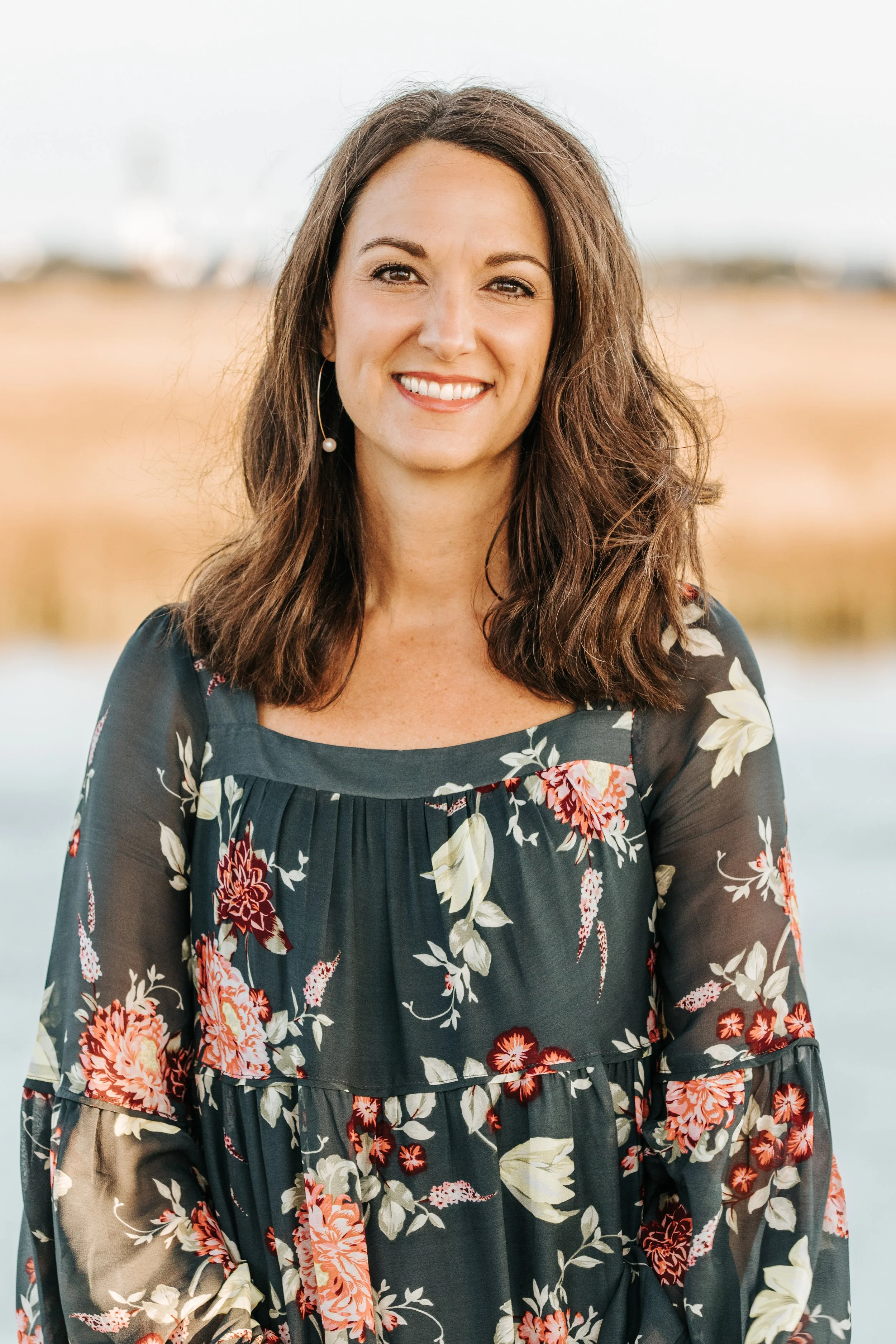 A woman with shoulder-length brown hair, smiling and wearing a floral dress, standing outdoors near water with blurred landscape in the background.