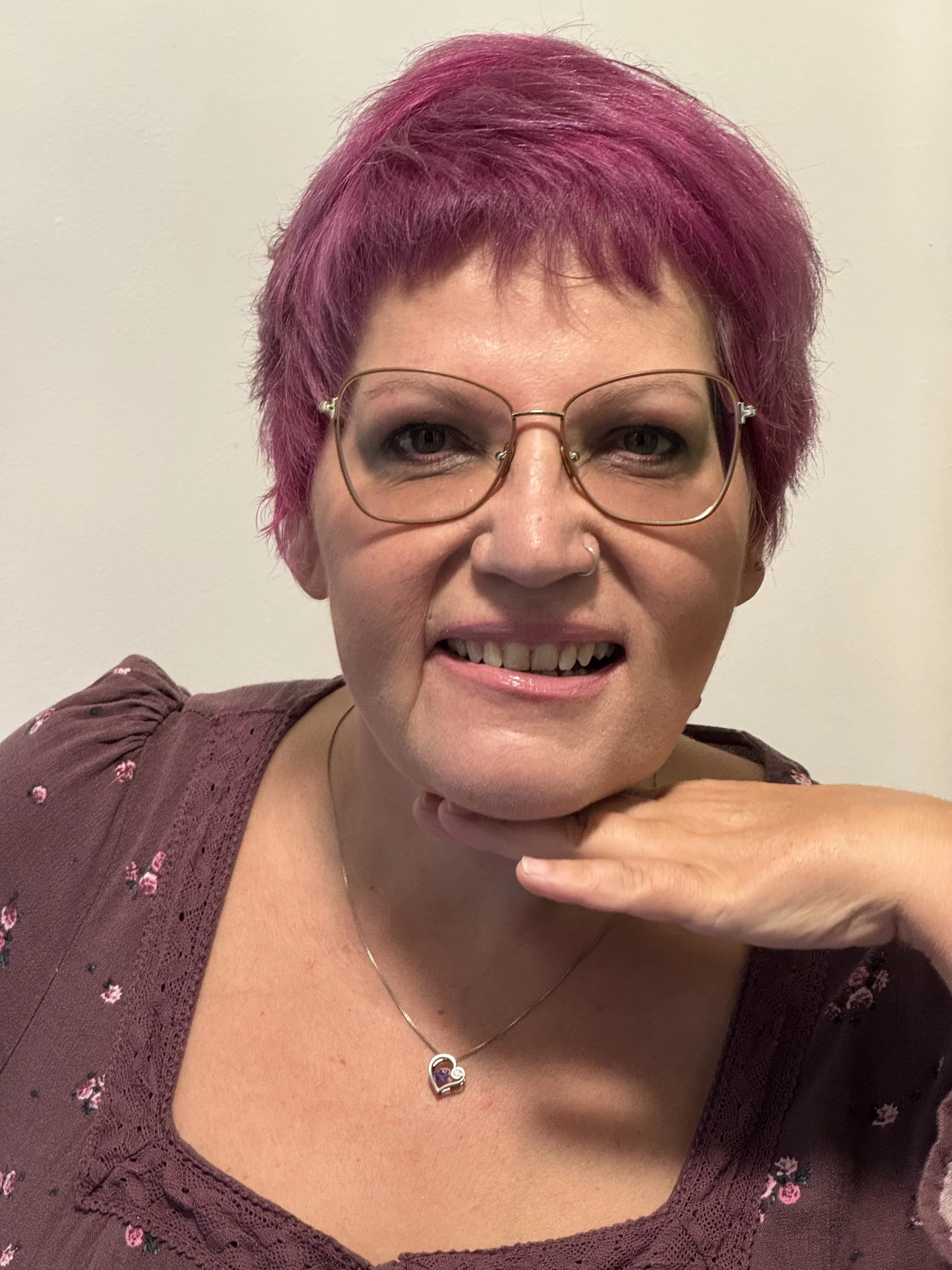 Close-up of a woman with short, vibrant pink hair, glasses, and wearing a maroon top with a floral pattern. She has a friendly expression and is resting her chin on her hand, showing a necklace with a heart-shaped pendant.
