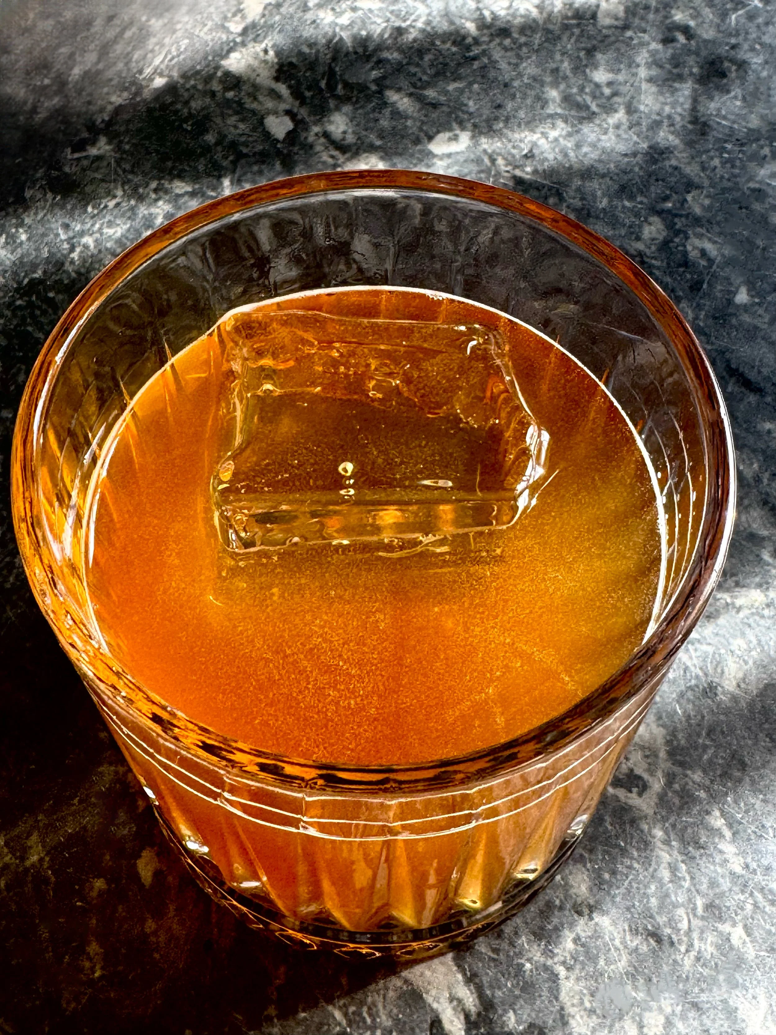 A glass of whiskey with a large ice cube on a dark textured surface.
