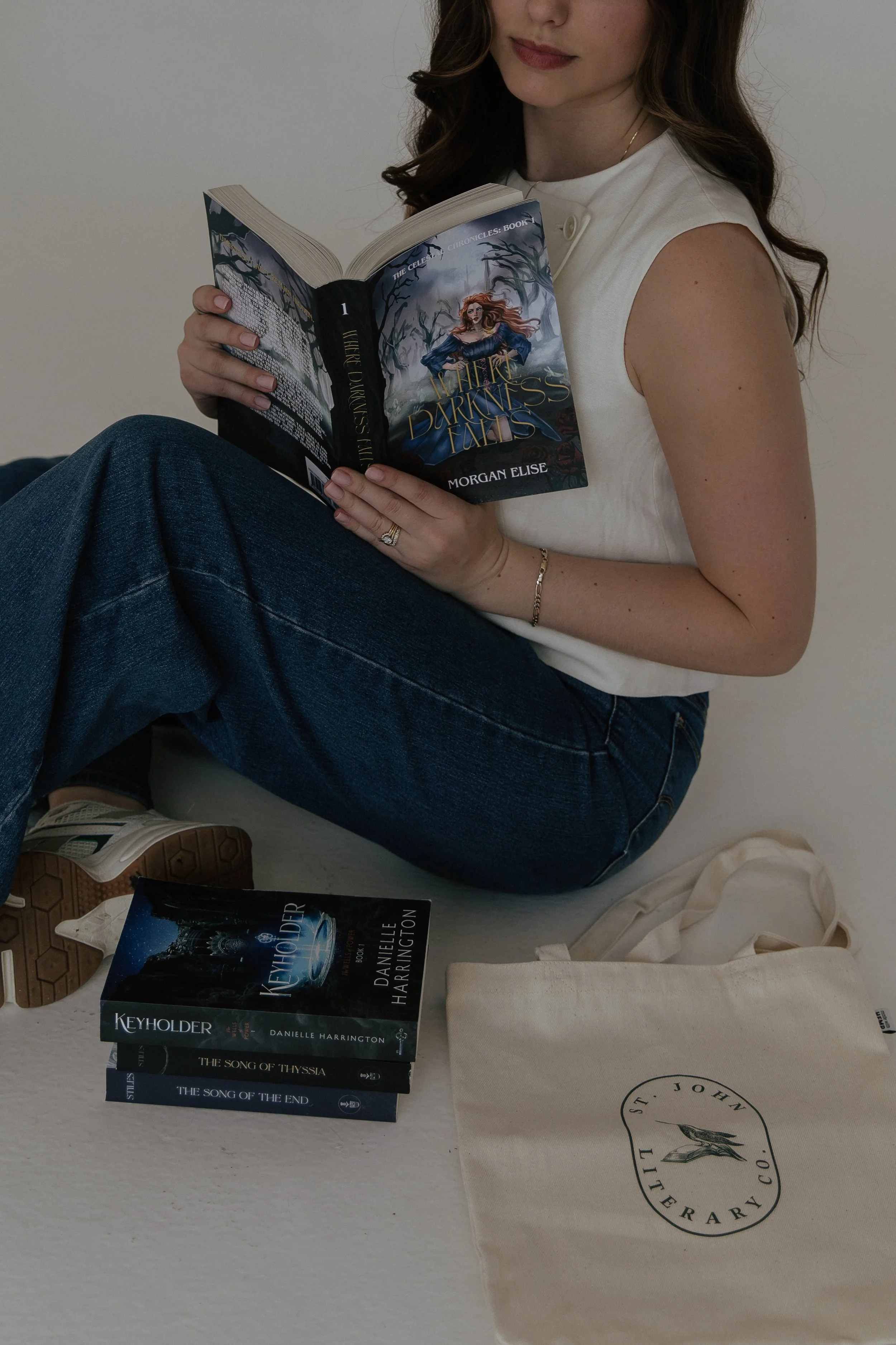 A woman sitting on the floor reading a book titled "The Darkness Falls." There are several stacked books including "Keyholder" and "The Song of Thyssia" nearby, and a tote bag with a circular logo for a bookstore.
