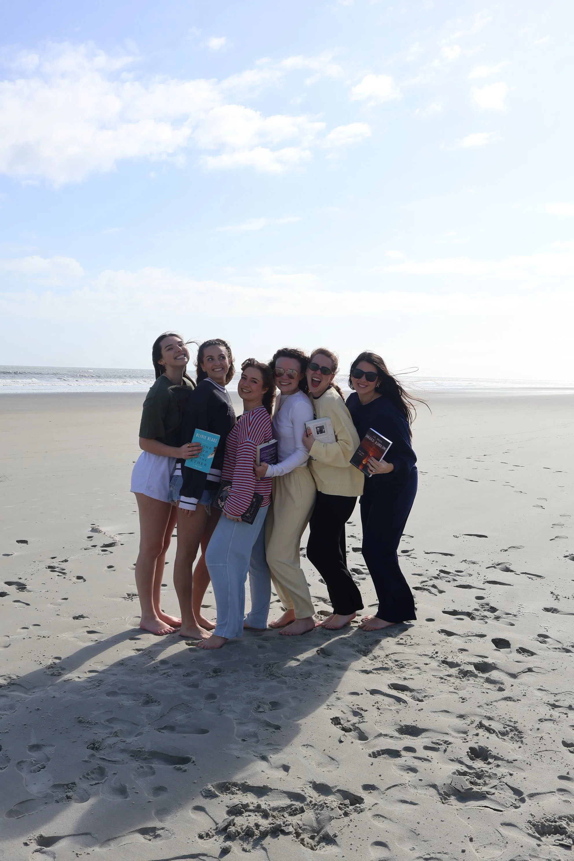 Group of seven women smiling and posing on a sandy beach with the ocean and sky in the background, holding books and standing barefoot.