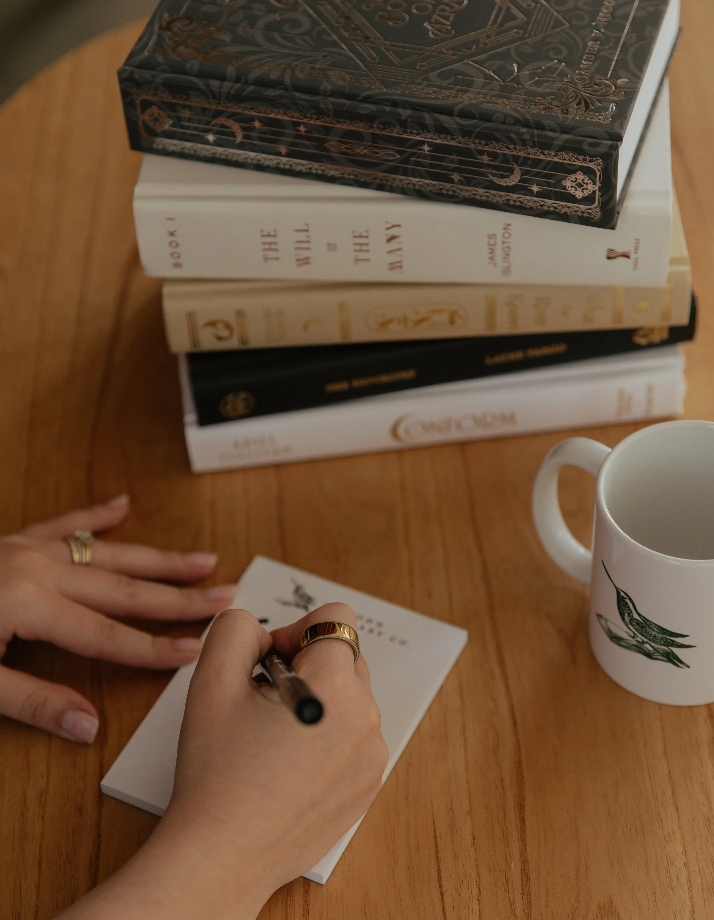 Stacked books on a wooden table with a person holding a pen and writing on a piece of paper, a white mug with green leaf design nearby.